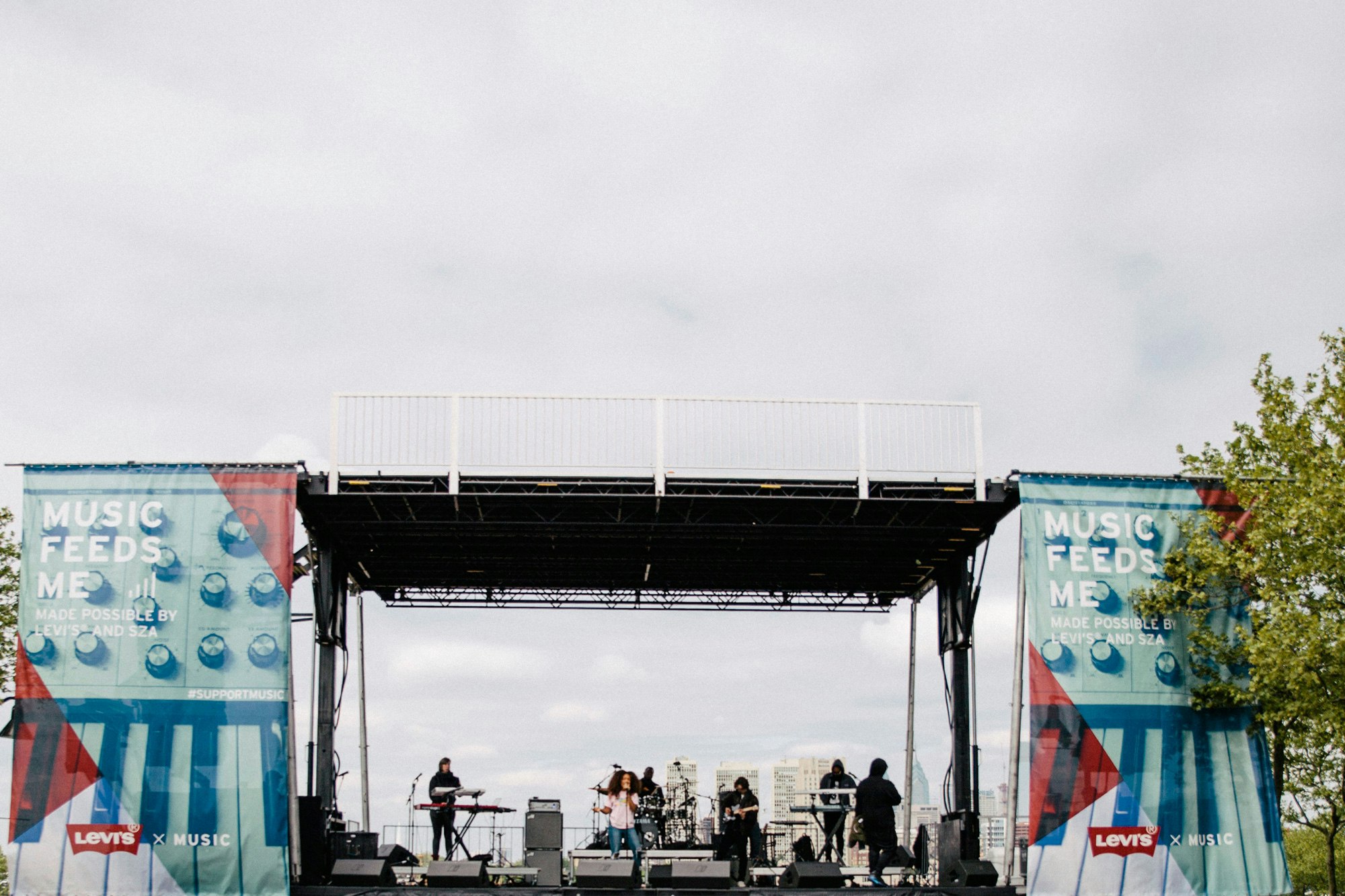 Image shows a large outdoor stage from far away. The sky is cloudy and there's a tree to the right of the stage. On either side of the stage hangs a banner that says MUSIC FEEDS ME on top of an image of a keyboard (instrument).