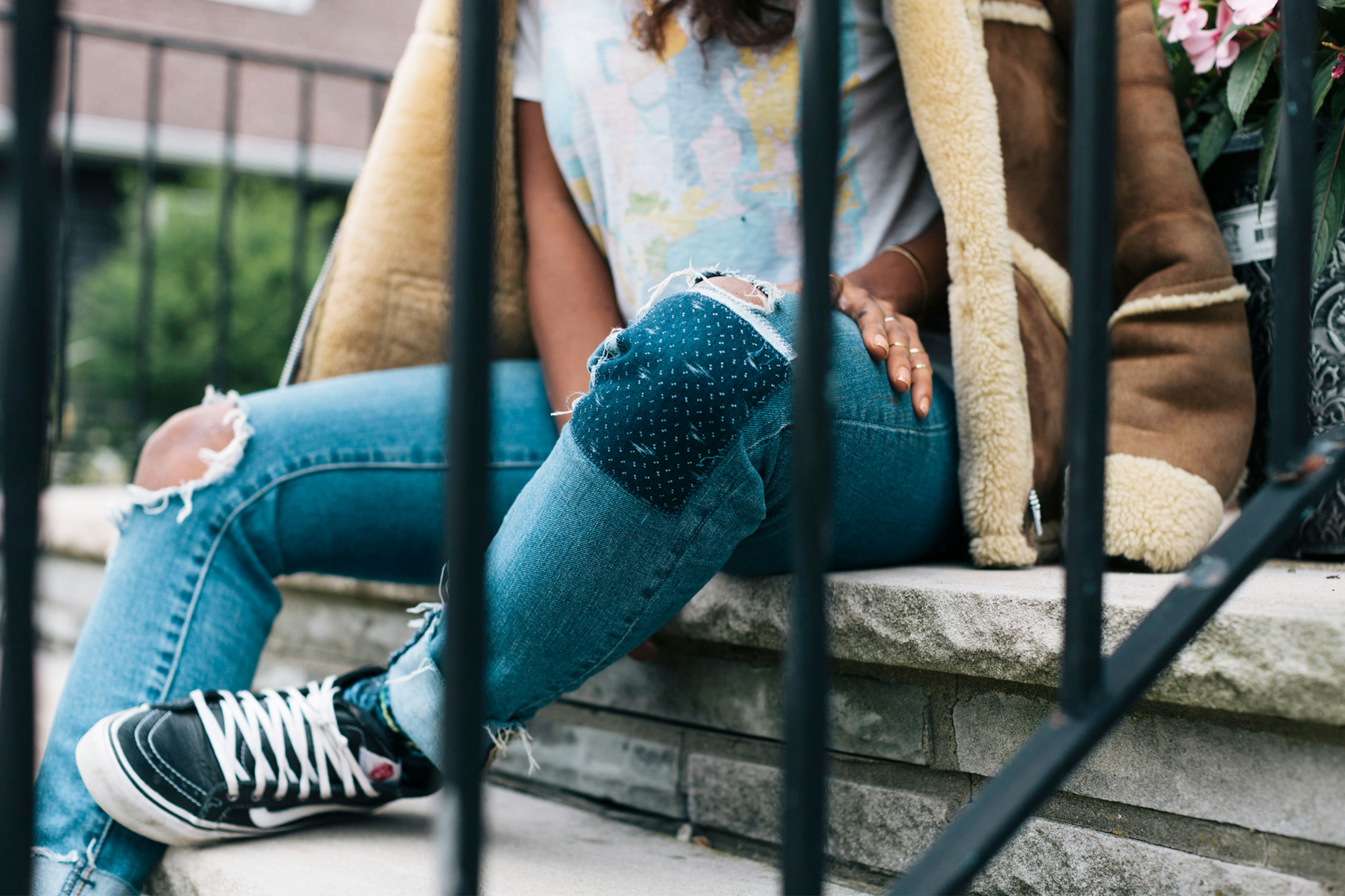Image shows a close up of a persons knee in ripped and patched blue jeans. They are also wearing black and white high top Vans sneakers and a tan sherpa lined jacket. 