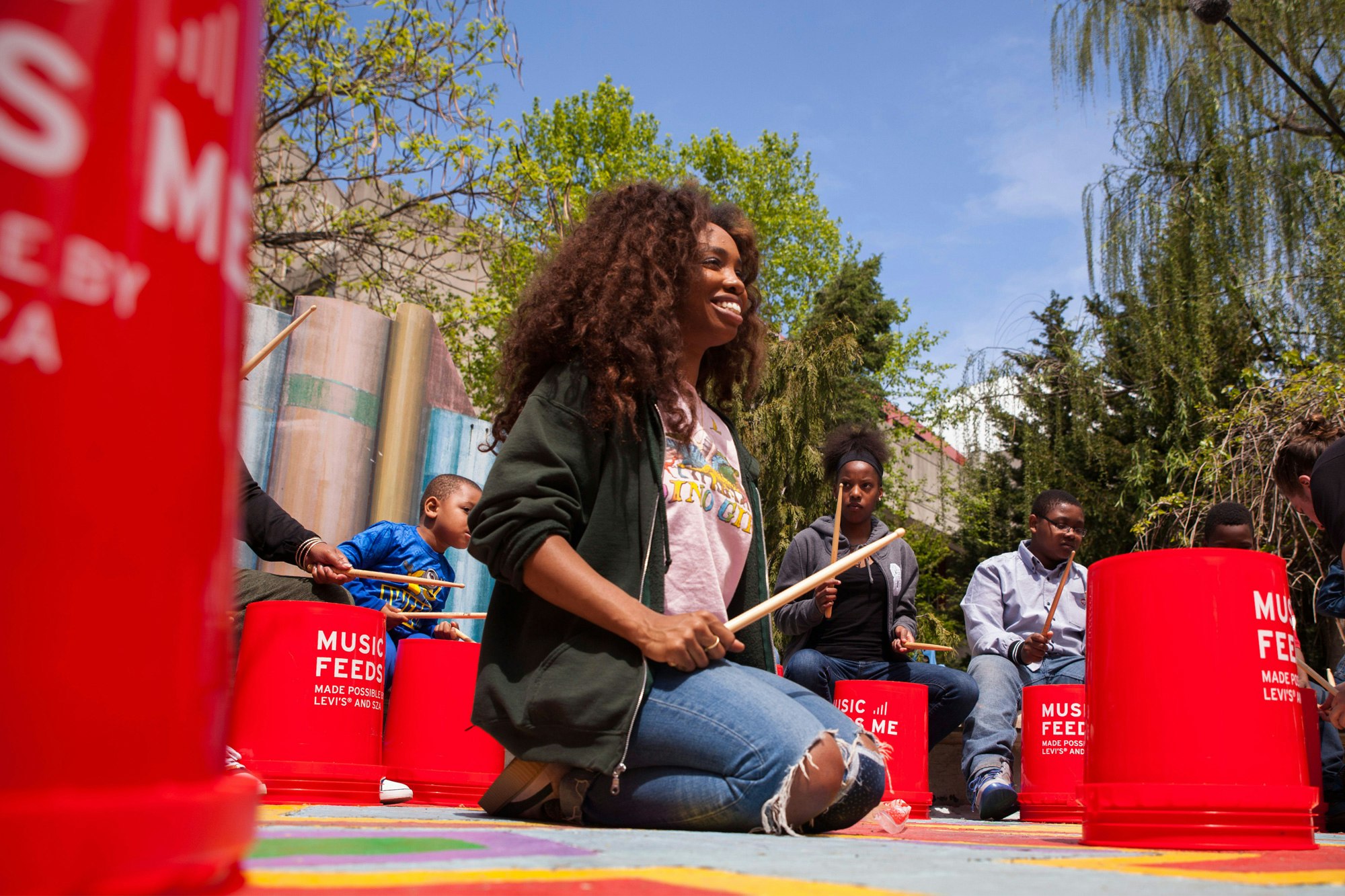 Image shows SZA outdoors, sitting in the middle of a circle of bright red buckets being used as drums. She is holding a drumstick and smiling. Behind the circle of drums and people is a blue sky and trees.