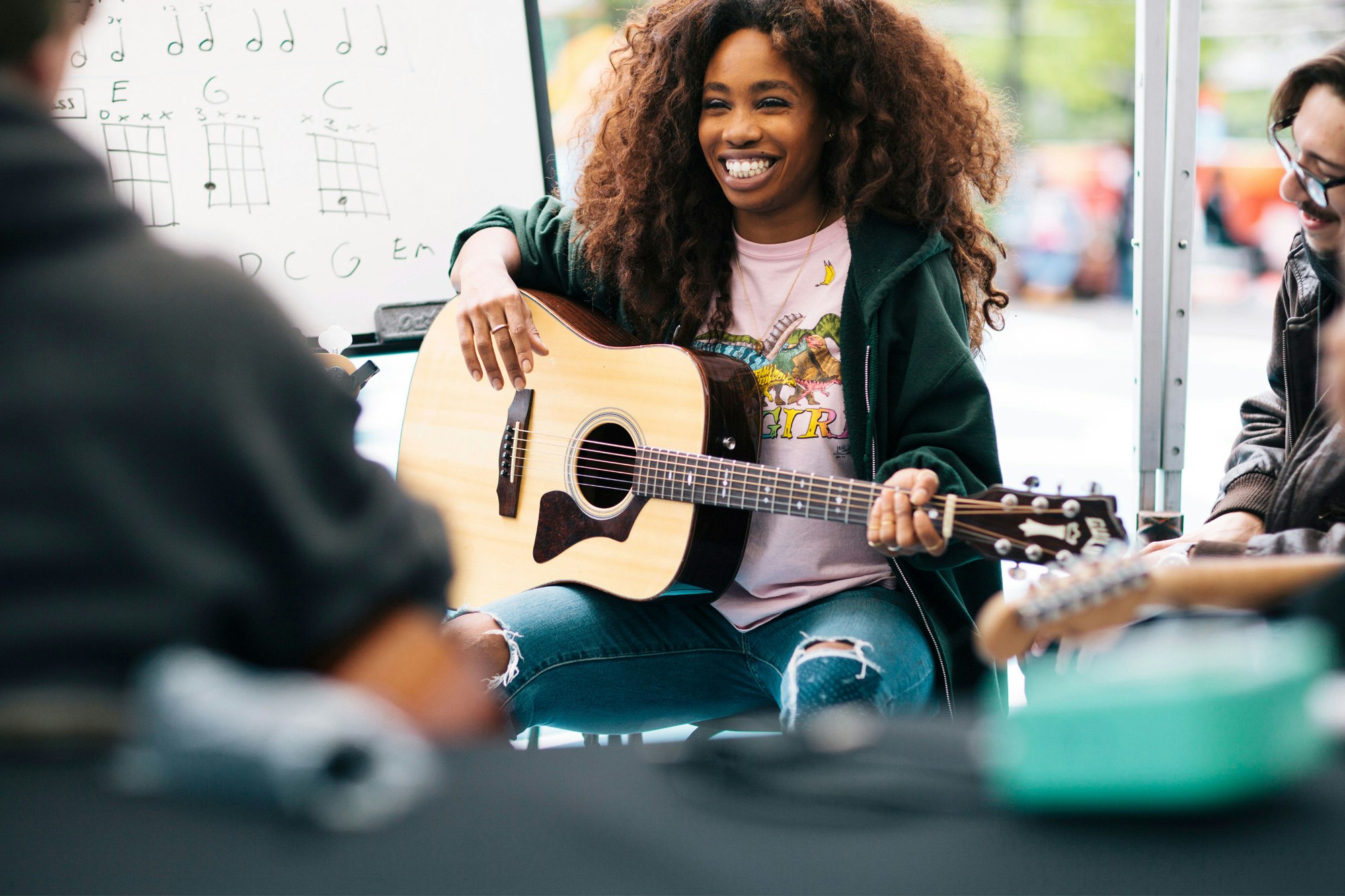 Image shows SZA sitting with an acoustic guitar. She is wearing a dark green hoodie and ripped jeans and smiling.
