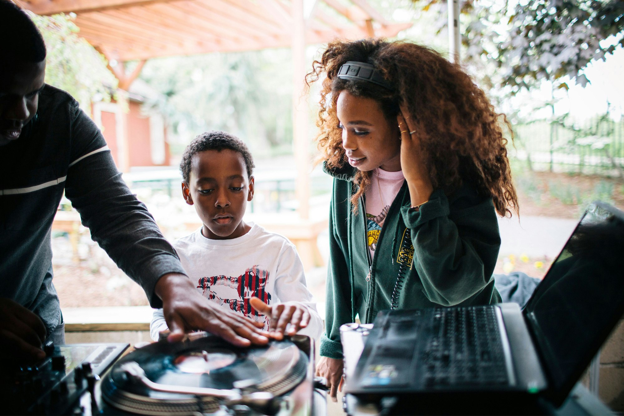 Image shows SZA wearing headphones, watching a child with their hand on a record. Another person stands to the left with their hand also on the record.