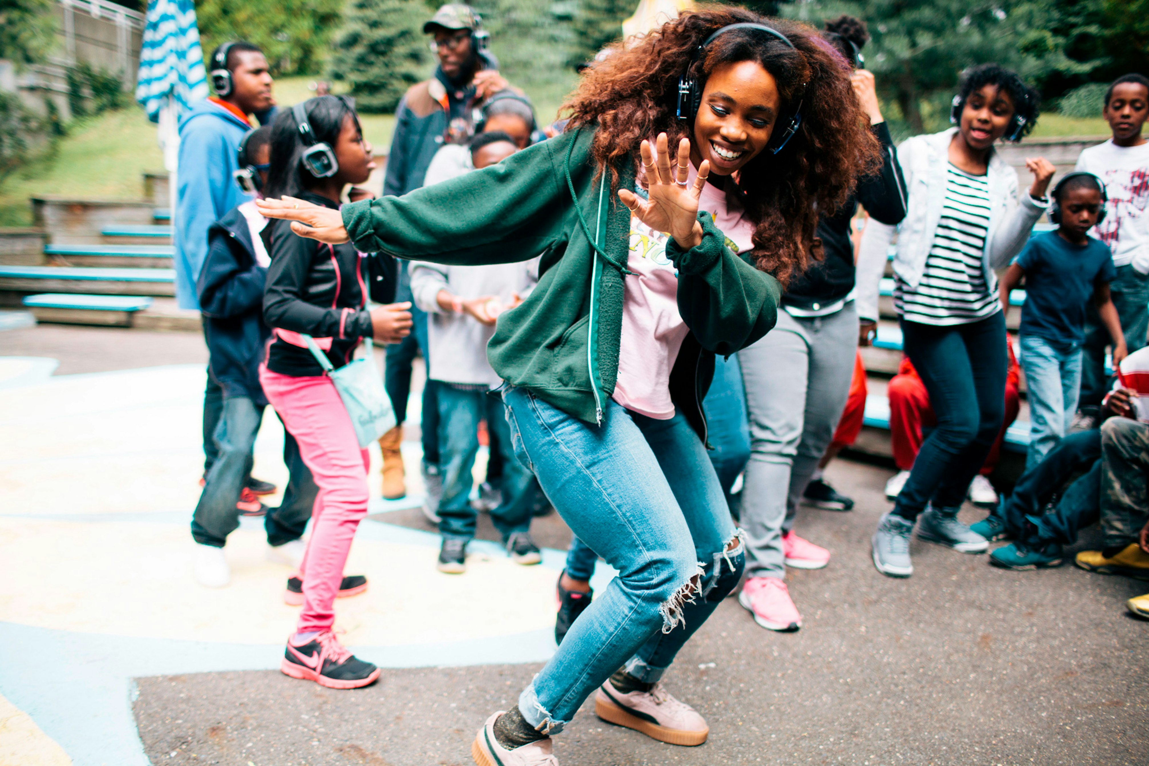 Image shows SZA wearing headphones, a green hoodie, and blue jeans dancing in front of a crowd of children and young people wearing headphones as well. They are outside and there are trees in the background. 