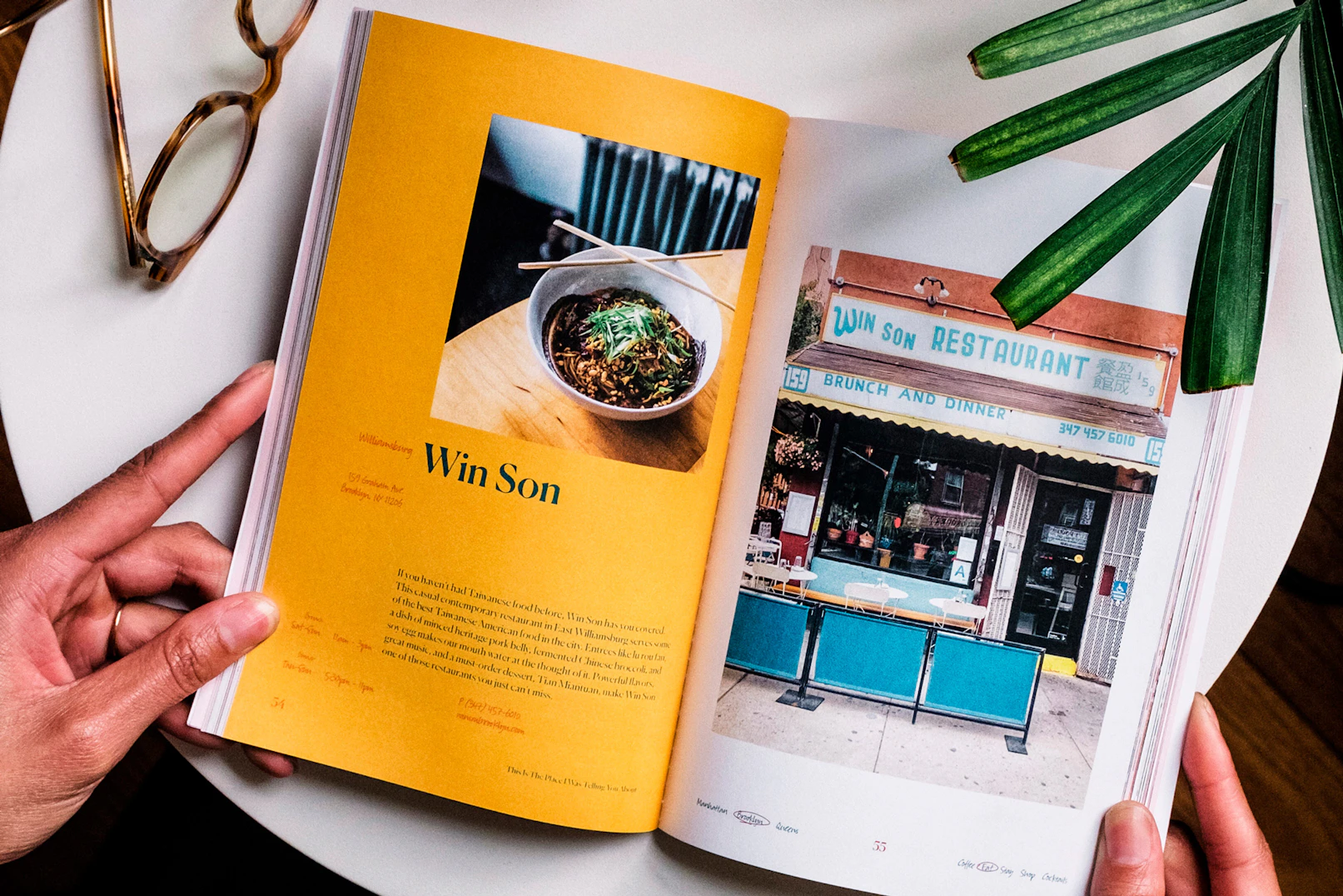 Image showing two hands holding the open book to a spread with a lot of yellow and blue colors. A pair of reading glasses are on the table and a segment of a palm leaf is coming into the image.