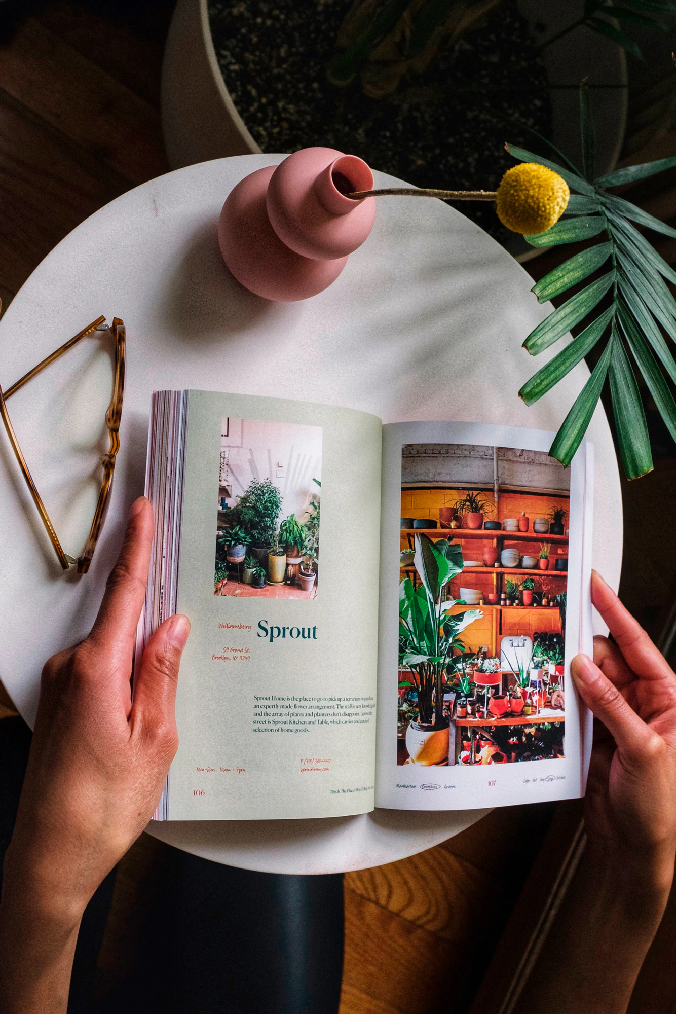 Hands holding the book open to a page showing pale mint color frame and photos from a plant store. On the white table there are reading glasses and a pink vase with a yellow flower.