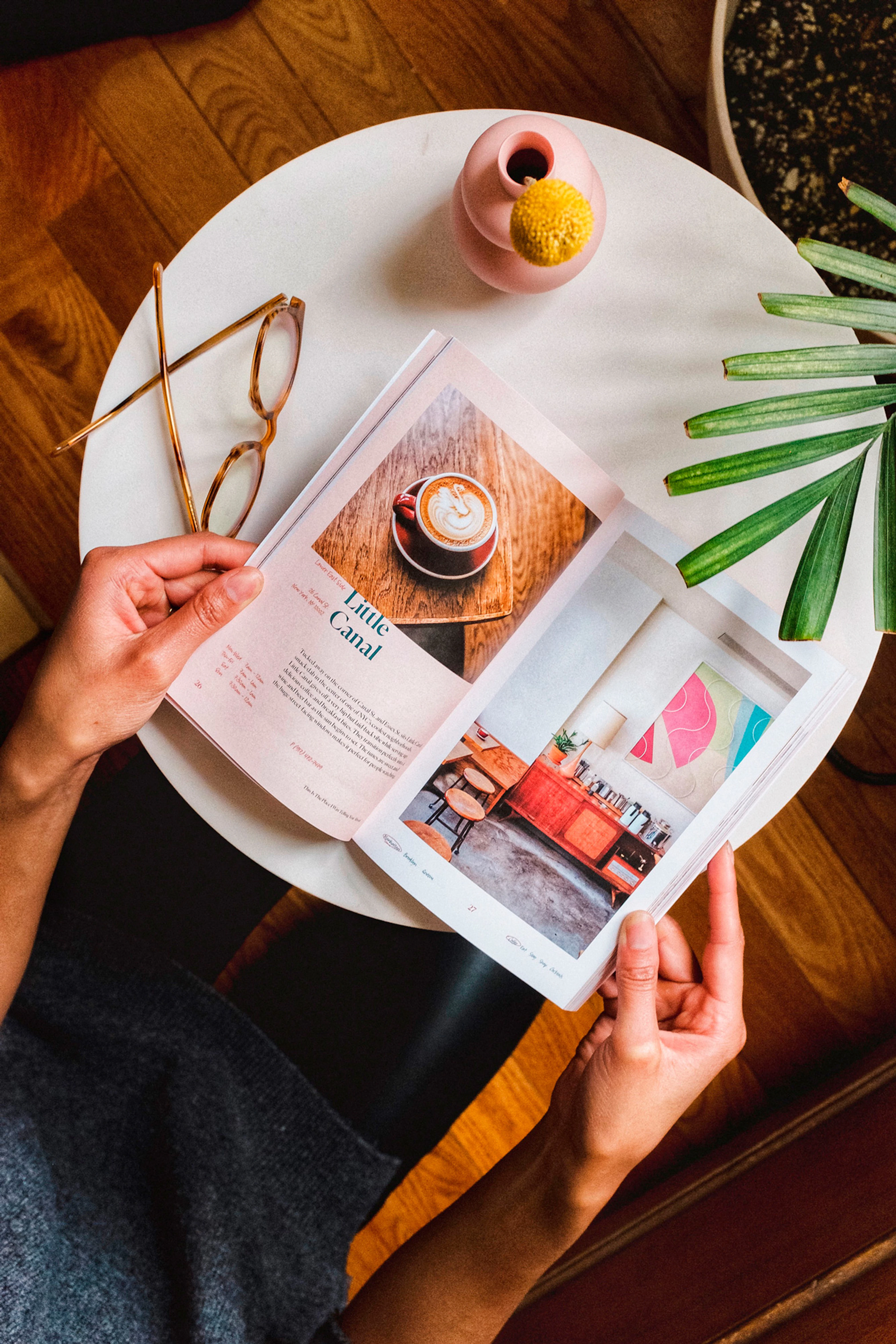 Hands holding the book open to a page showing a cup of cappuccino and an indoor space with an orange couch and a large piece of modern art hanging on the wall. 