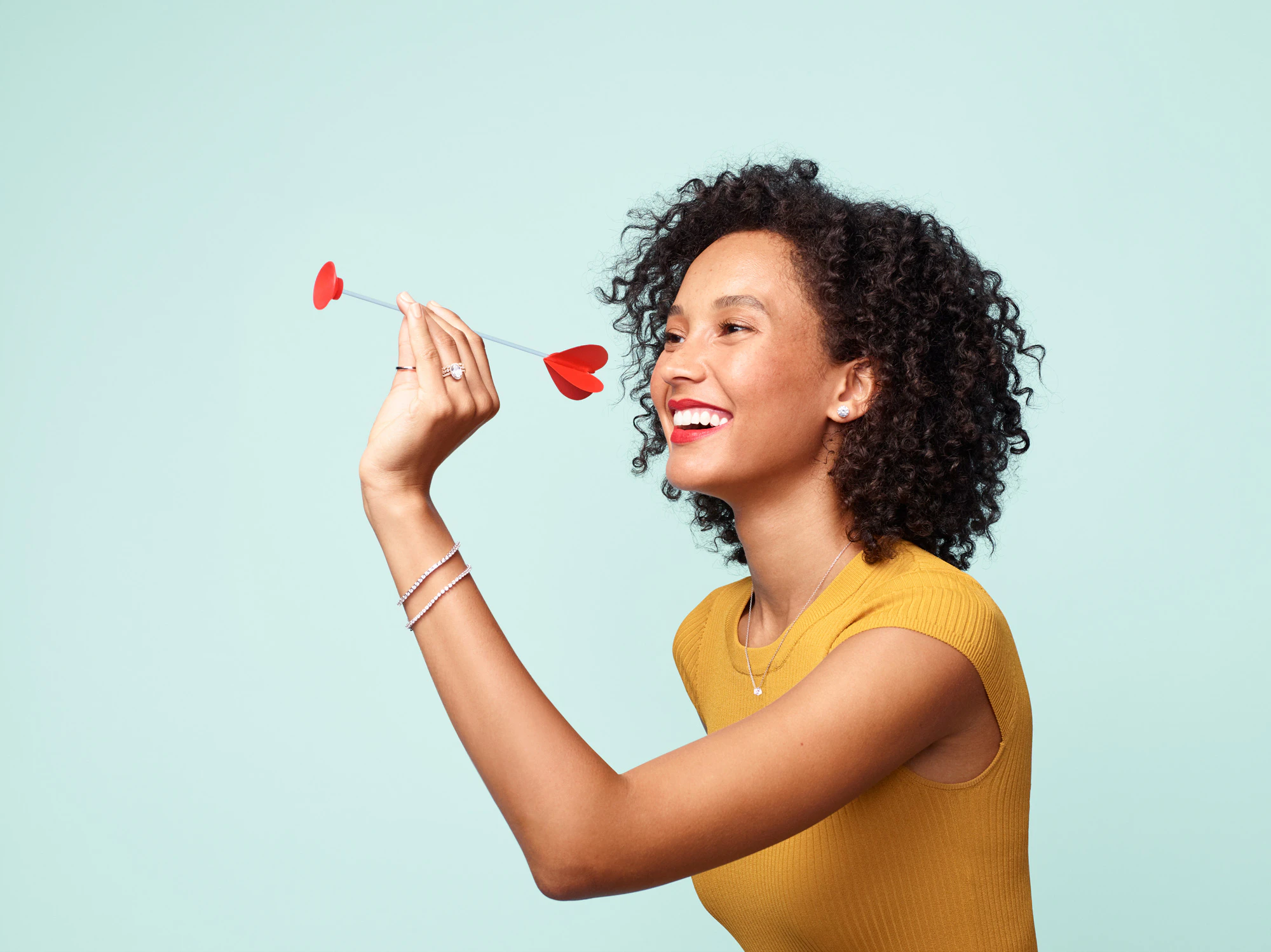 Image showing person holding a red heart arrow ready to send it to their target. The person is wearing a mustard yellow top and the seamless is frosted light blue.