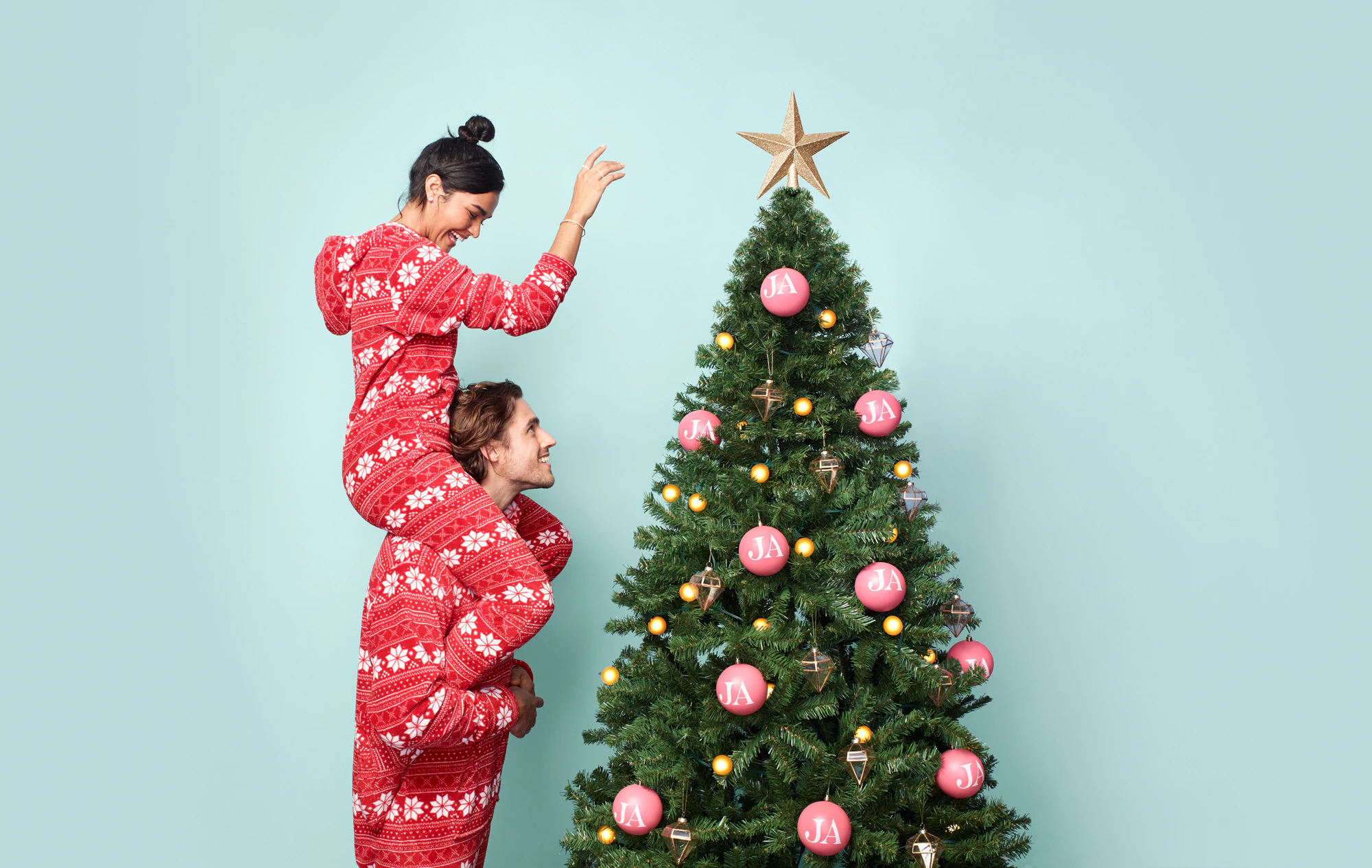 Image showing two people, one on the other persons shoulders. They are wearing matching Christmas pajamas and they are putting the star on the Christmas tree.