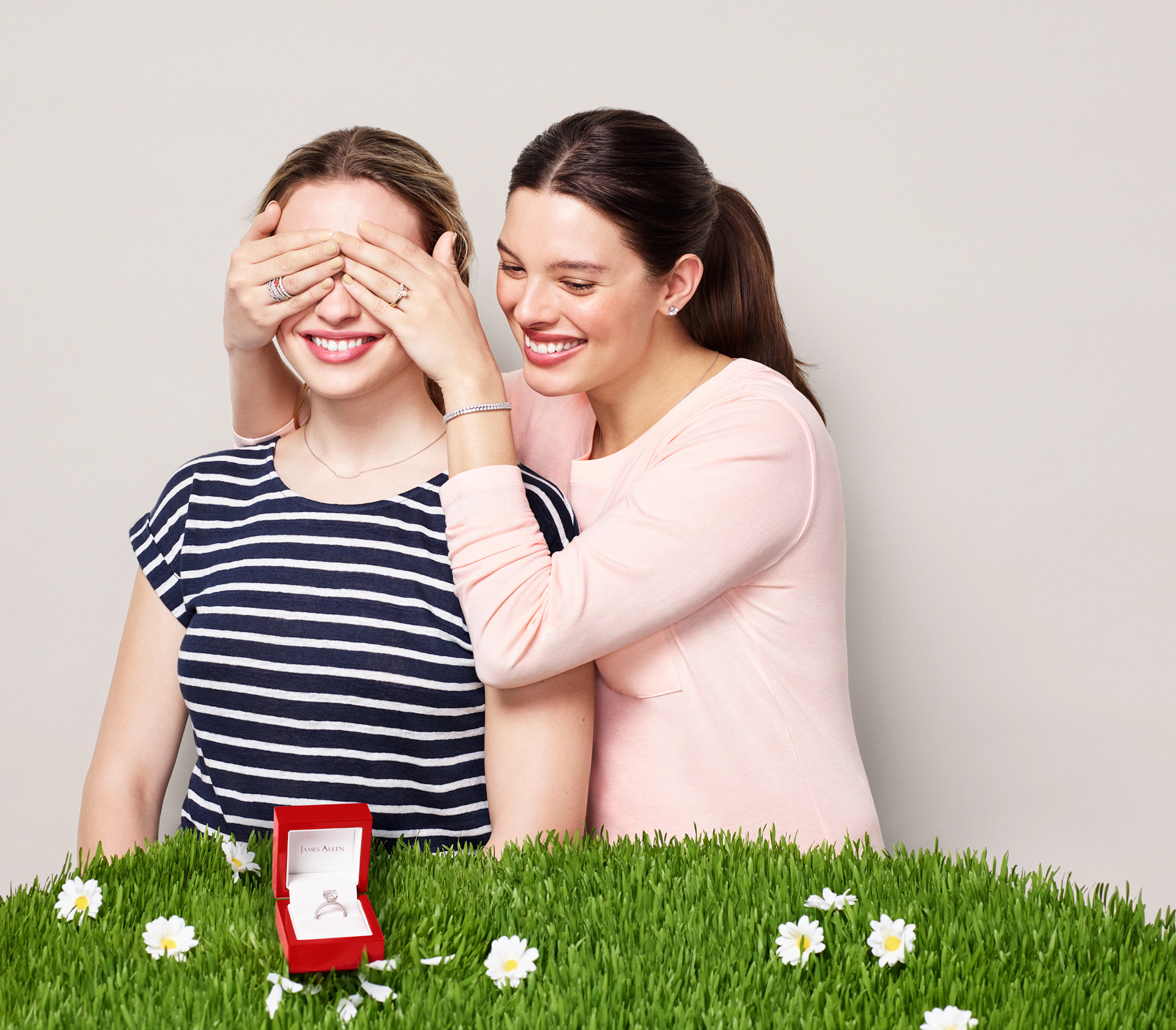 Two women are standing in front of a gray seamless and in front of them is grass with daisies. On top of the grass is an open JamesAllen ring-box with an engagement ring inside. One woman is covering the other woman's eyes with her hands and they are both smiling.