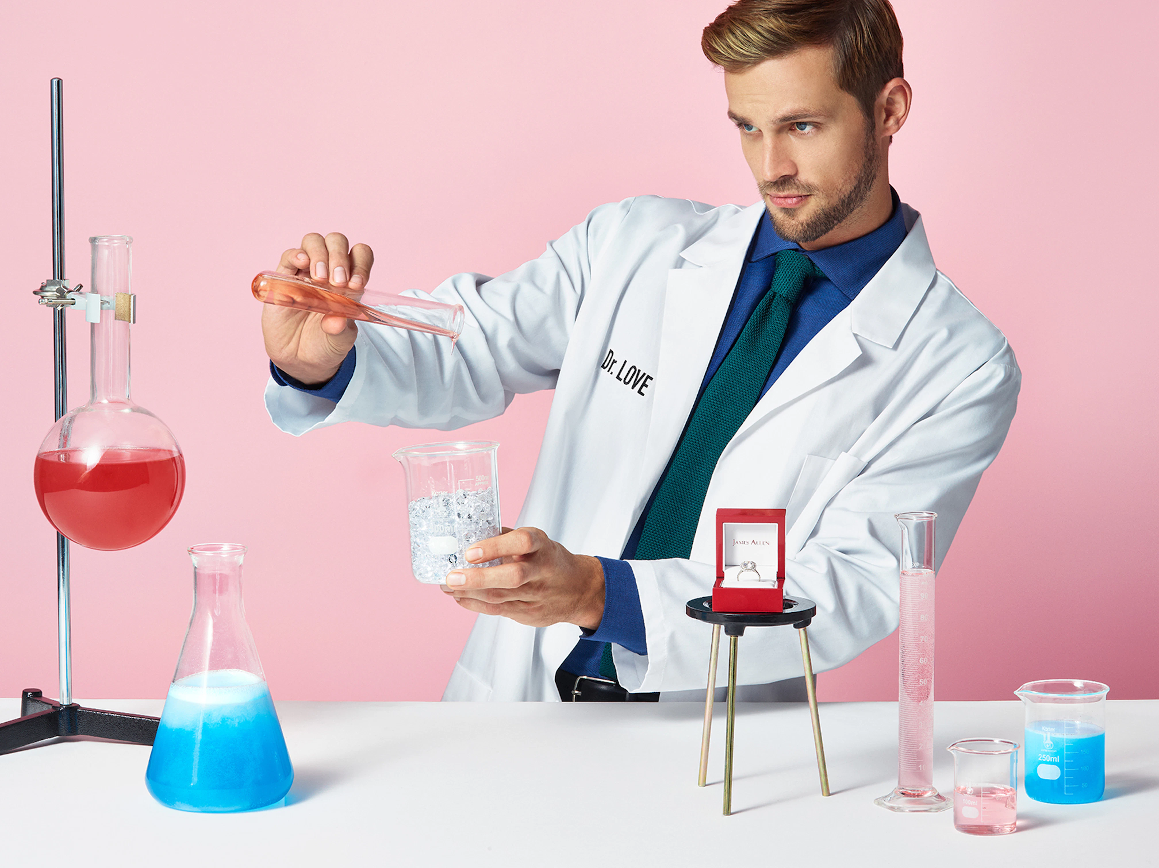 Image showing person in a chemistry lab. They are wearing a lab coat saying 'Dr Love' and on the table there is an open ring box with an engagement ring inside.