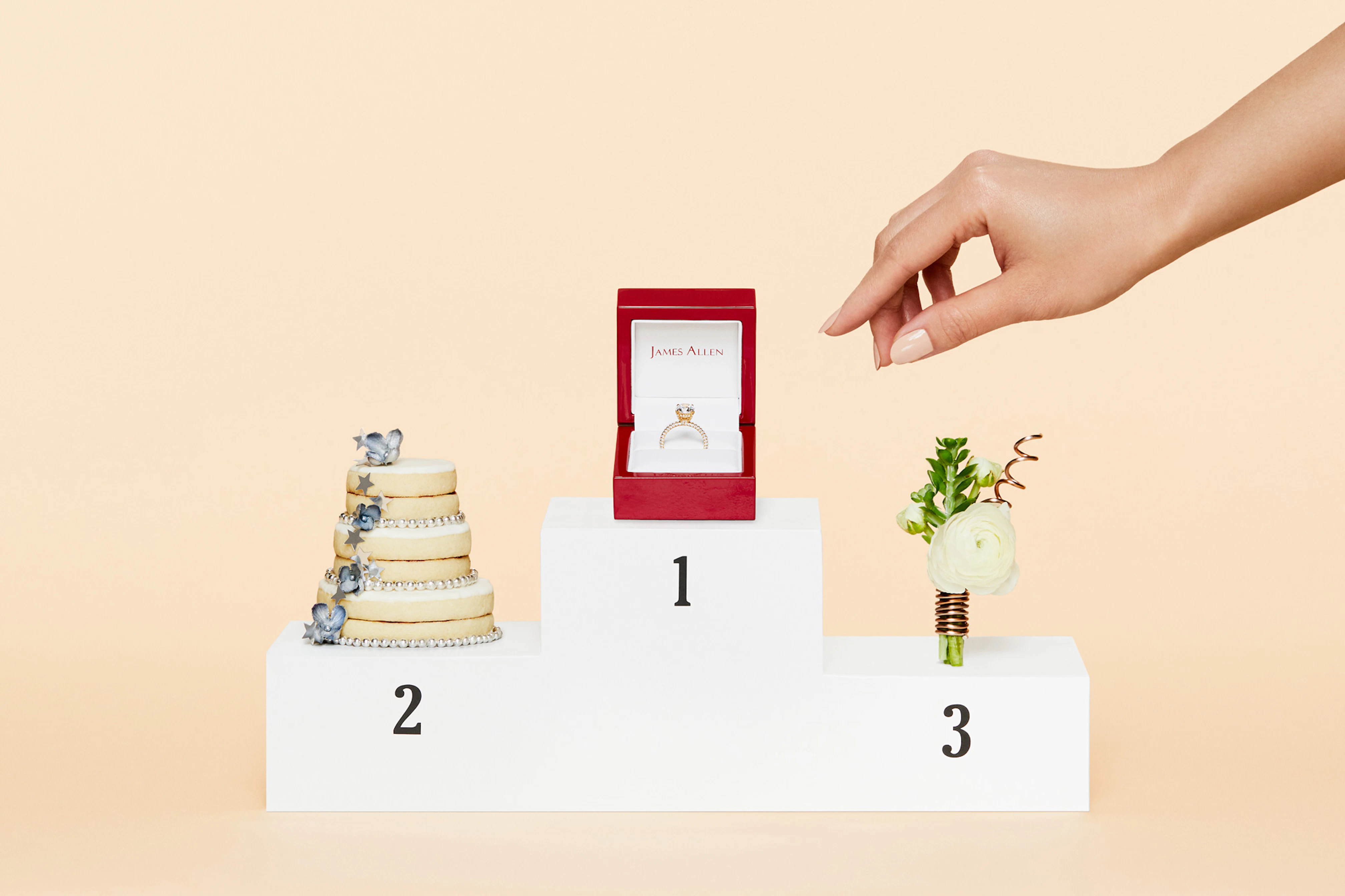 Image showing an Olympic winners steps with 1, 2, 3 written on the white steps in black. On the third place is a mini bouquet of flowers wrapped in bronze metal wire. On the second place is a mini wedding cake decorated in silver and on the first place is an open James Allen ring box with a red gold engagement ring inside. A hand is reaching for the ring box.