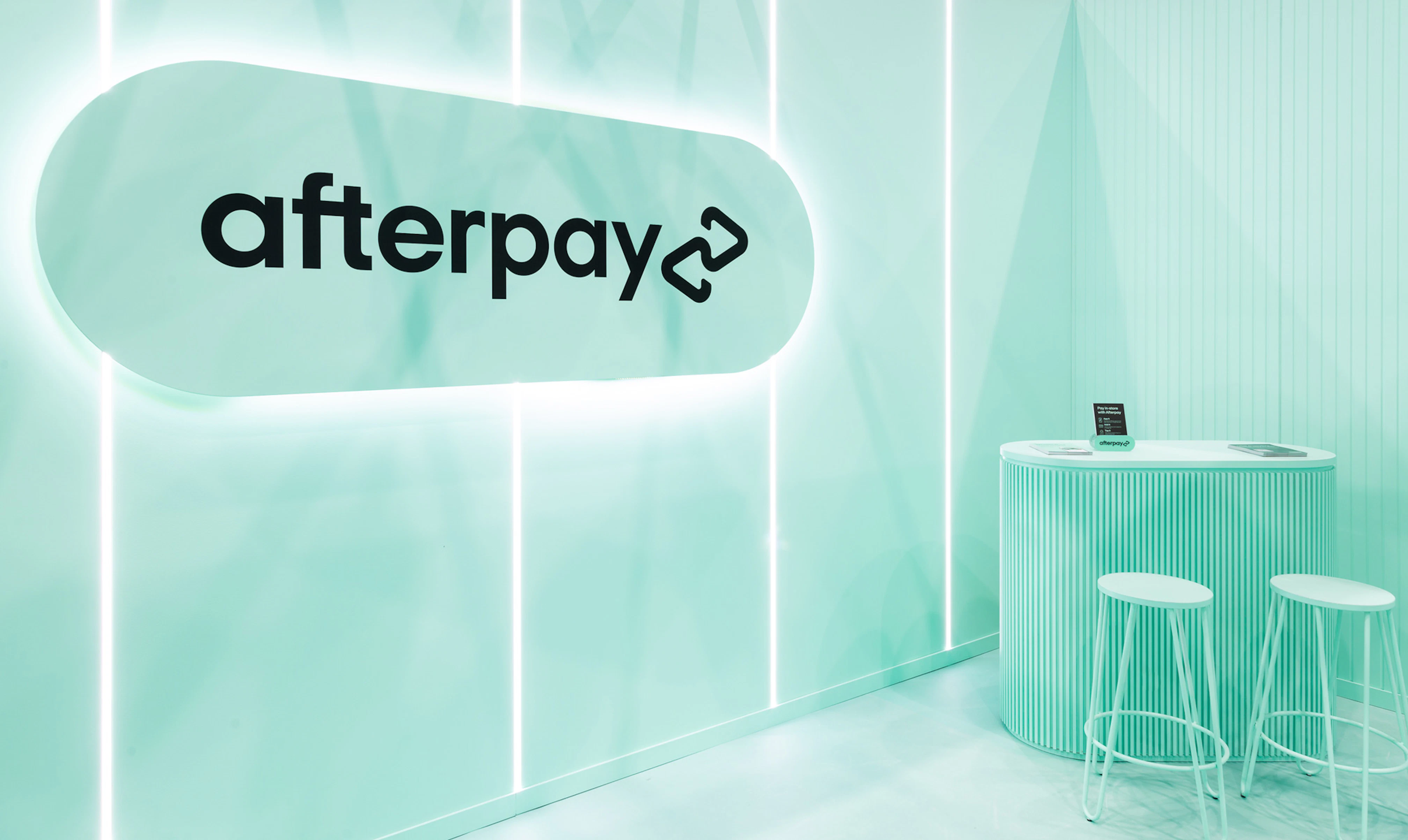 Image showing the interior of the Afterpay expo booth. Everything is Bondi Mint and there is an oversized logo pill which is backlit. In the back is a mint table and two mint stools. On top of the table are black pamphlets describing Afterpay.