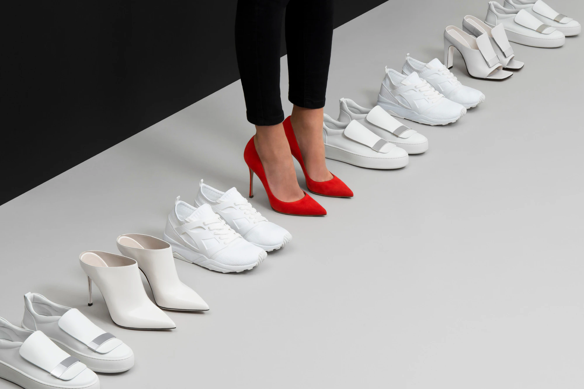 Image shows a line of white shoes with a person wearing one pair in the middle of the line, which is a pair of bright red heels.
