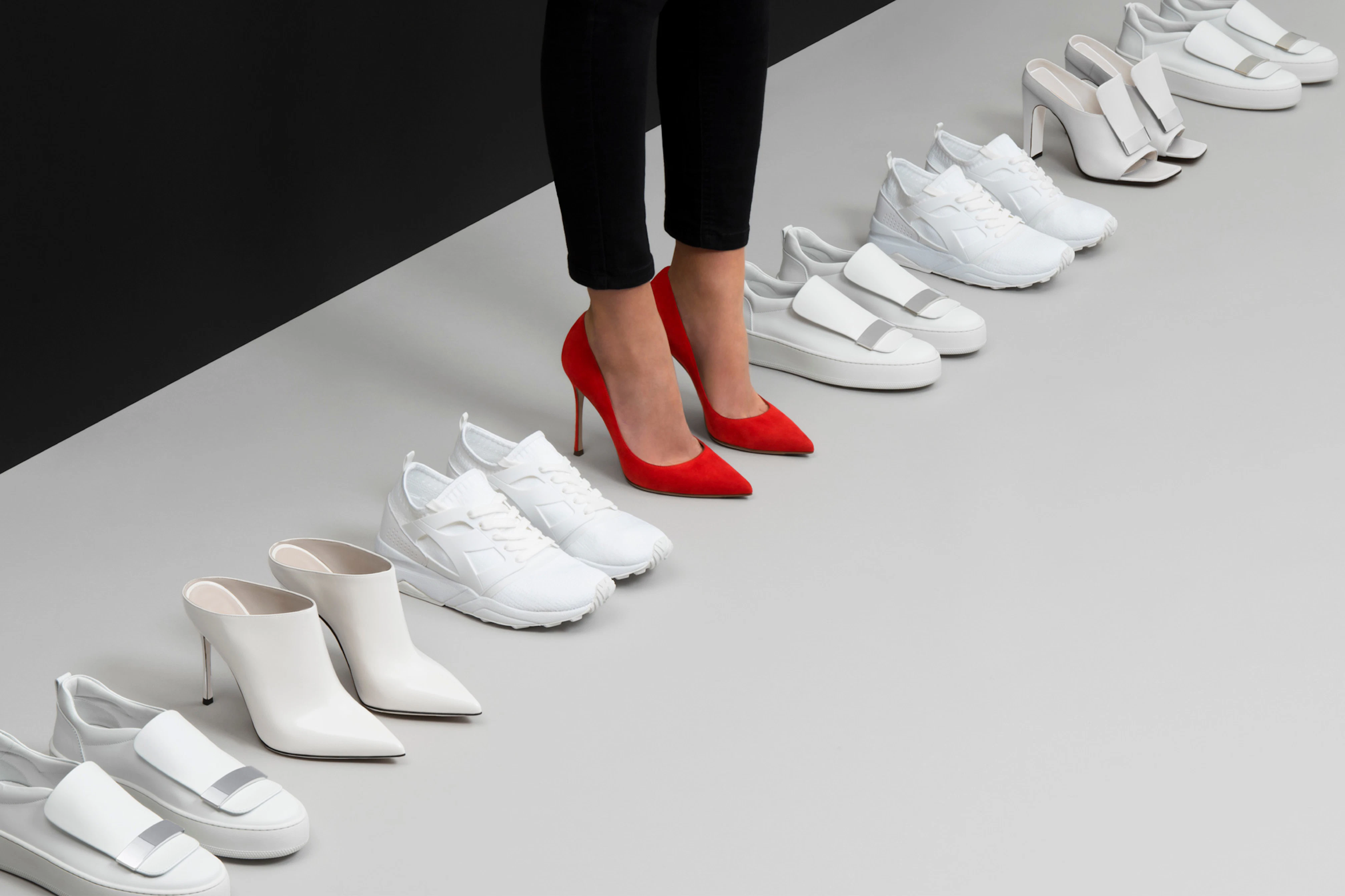 Image shows a line of white shoes with a person wearing one pair in the middle of the line, which is a pair of bright red heels. 