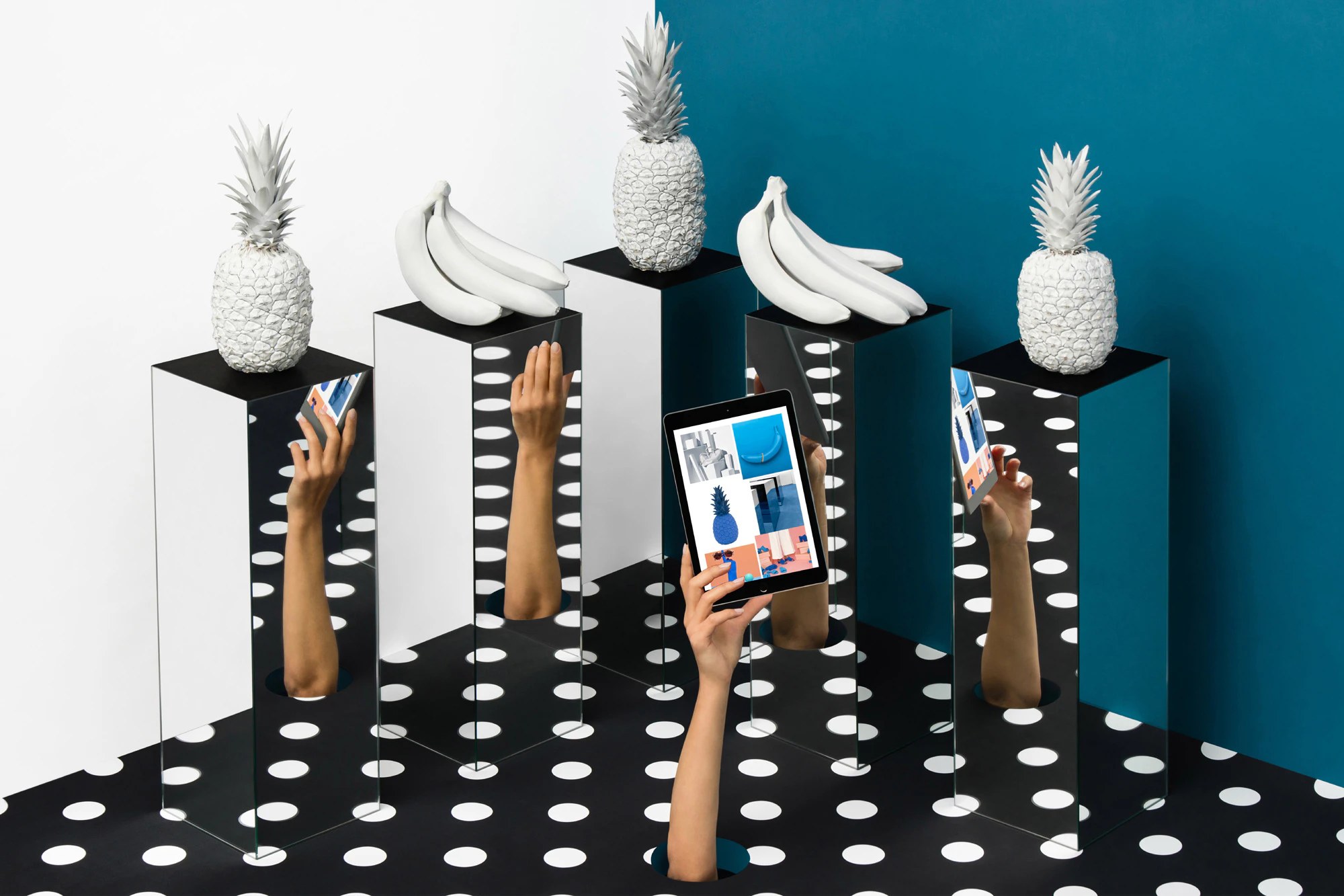 Image shows a hand reaching up through a black and white polka dotted floor holding a table. The hand is reflected on four mirror-surfaced pedestals with white fruit on top of them.