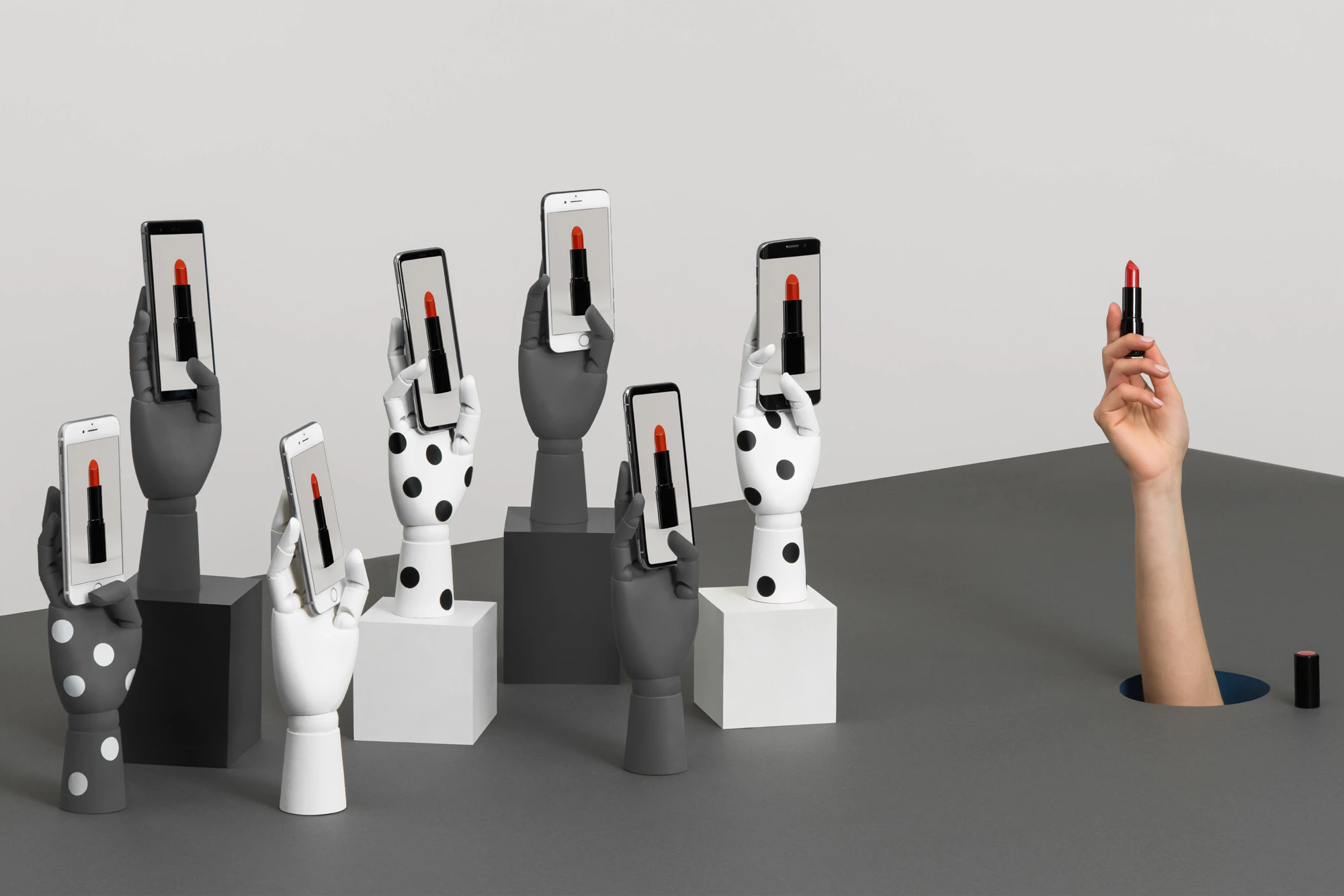 Image shows a hand reaching up through a hole in the gray floor holding a tube of lipstick. The arm is surrounded by a cluster of gray and white prop hands holding phones that display an image of the lipstick.