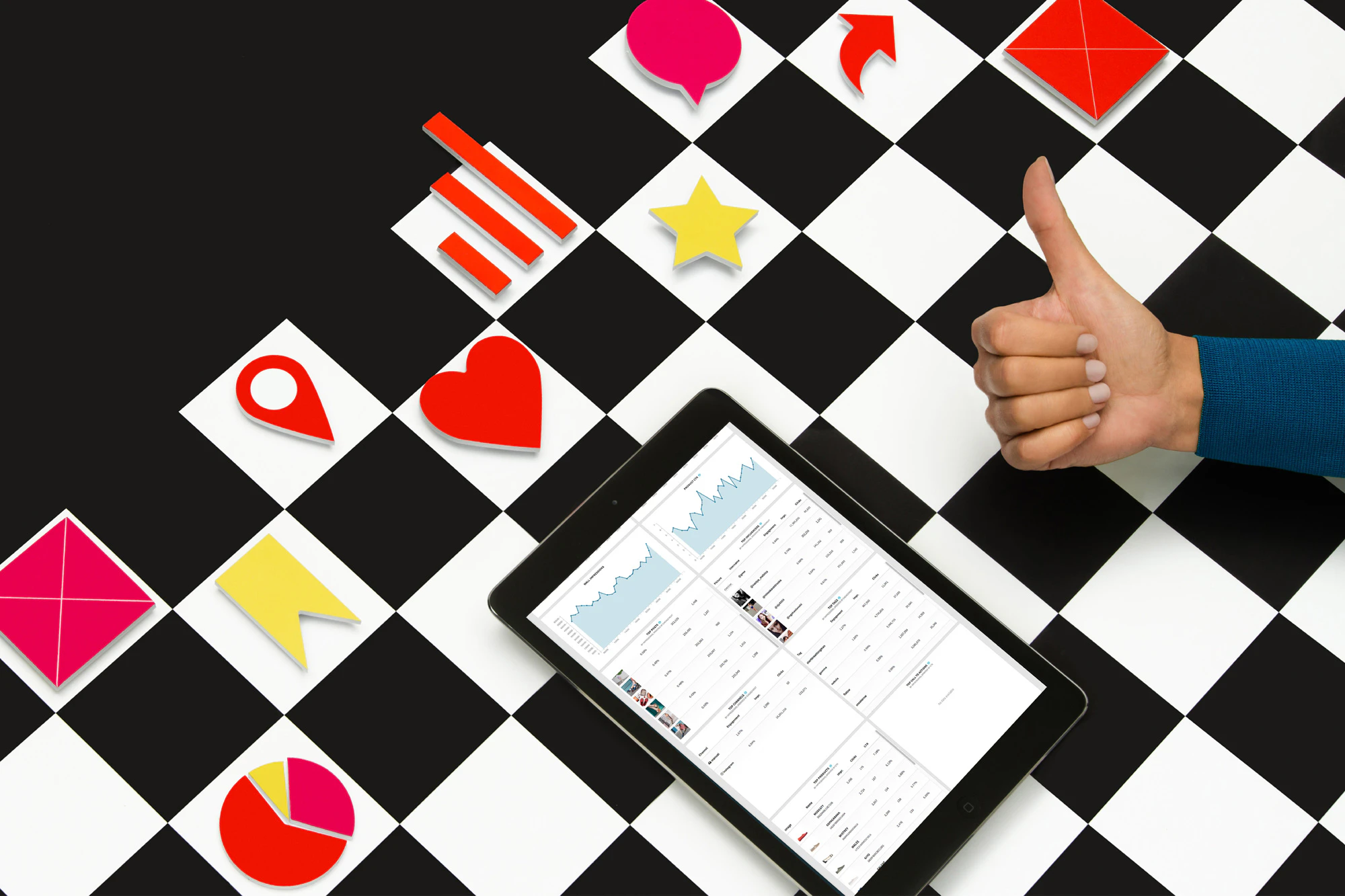Image shows a checkered surface with small icon pieces on it (an arrow, a pie chart, a star, a heart, a location pin, etc.). A tablet with data on it also sits on the board and an arm with a thumbs up hovers over it.