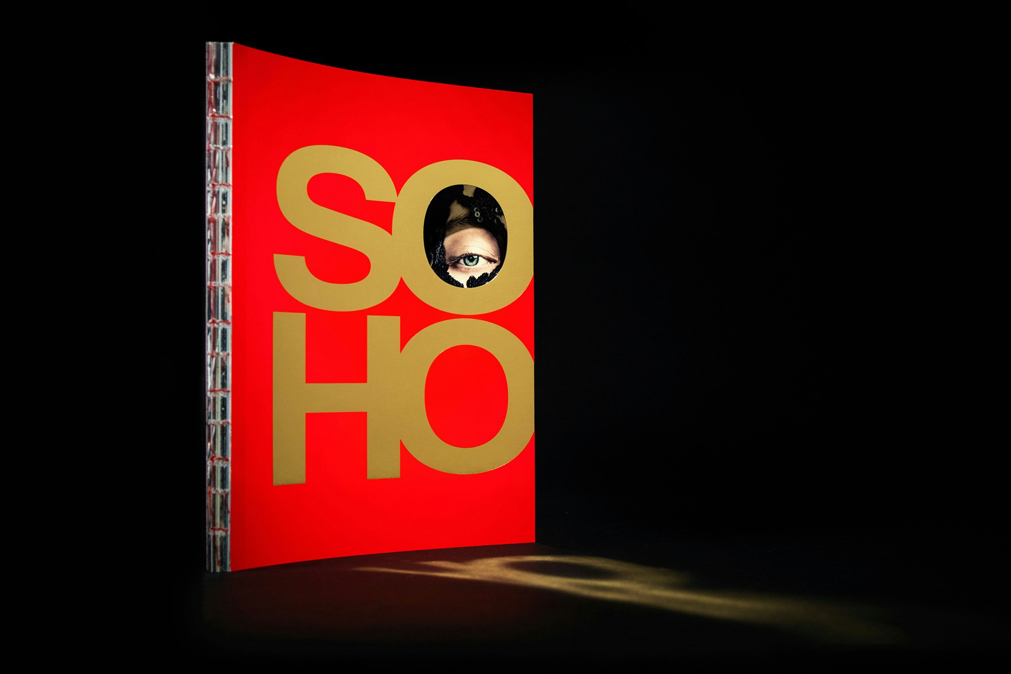 Image shows the book with the red cover and the gold letters SOHO and with an eye peeking through the first O standing on a black background, casting a gold reflection on the ground.