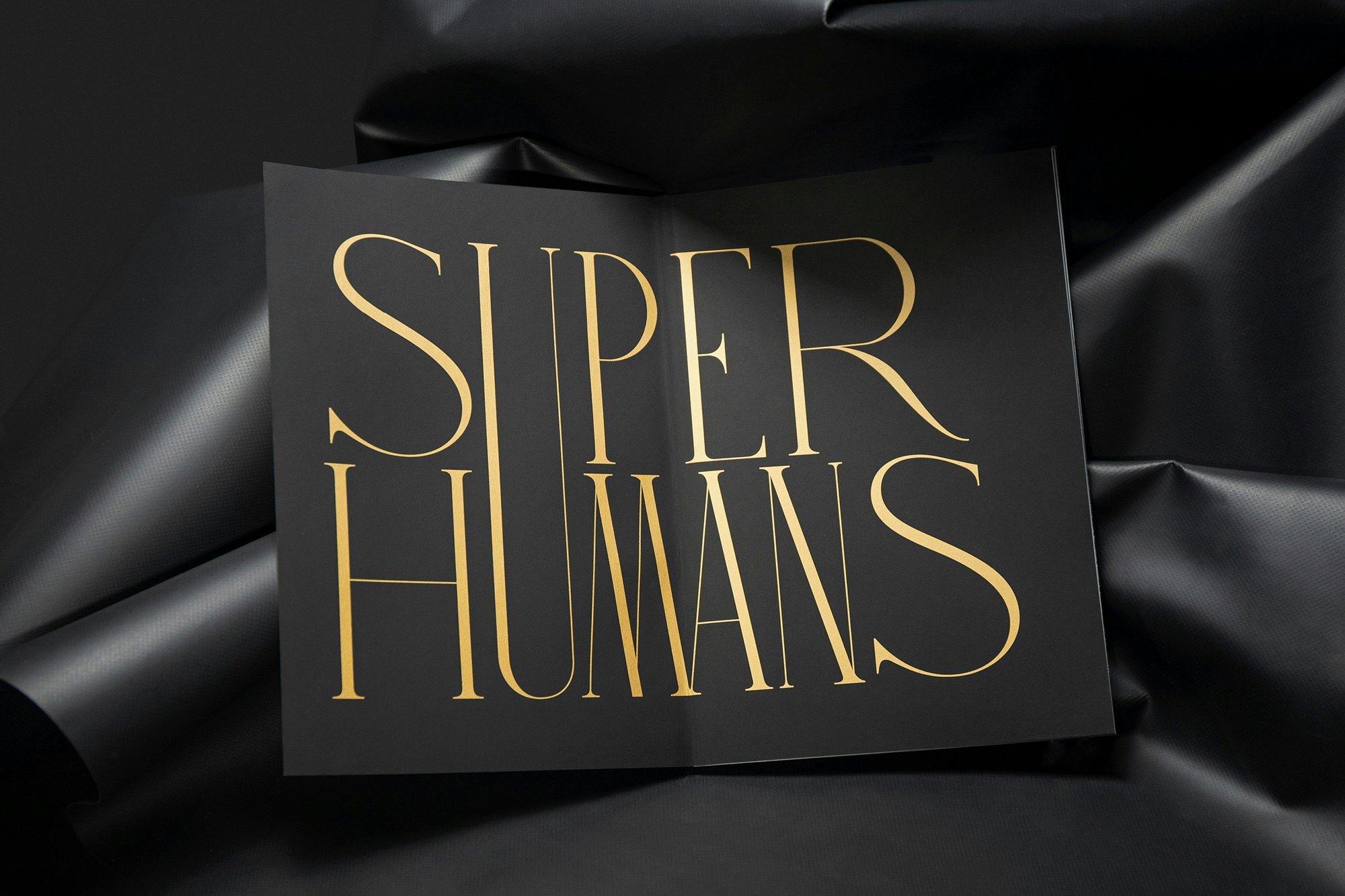 Image shows a book open to black pages with gold text reading SUPER HUMANS on it, with the U spanning both words. The book sits on a leathery black background.