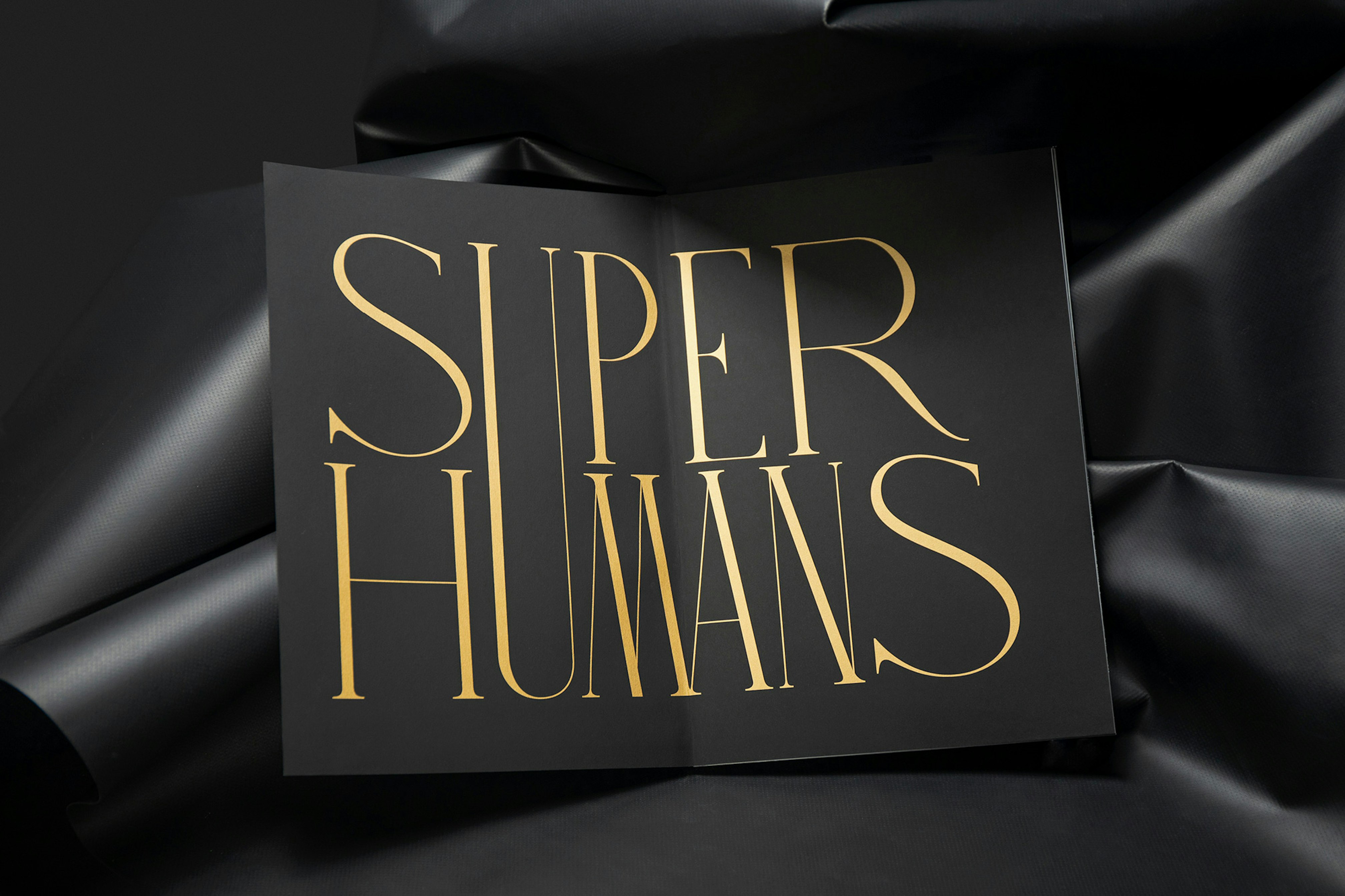 Image shows a book open to black pages with gold text reading SUPER HUMANS on it, with the U spanning both words. The book sits on a leathery black background. 