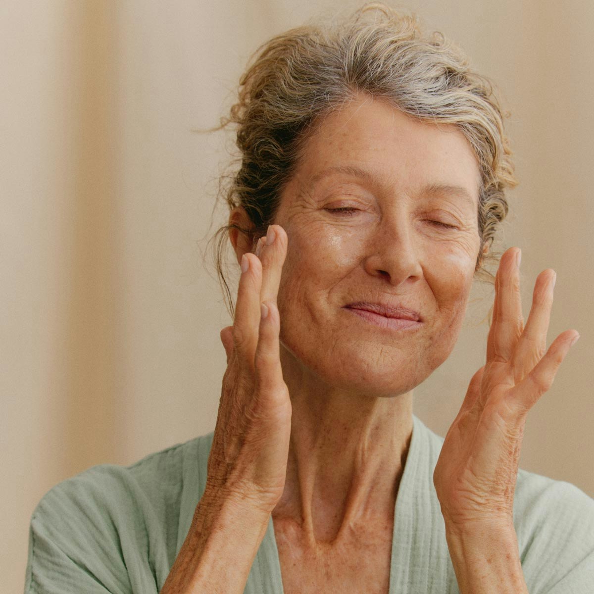 A photo of a radiant old person, eyes closed, wavy hair tied back. They are wearing a soft green robe and holding their hands up close to their face. They are smiling, looking joyful.
