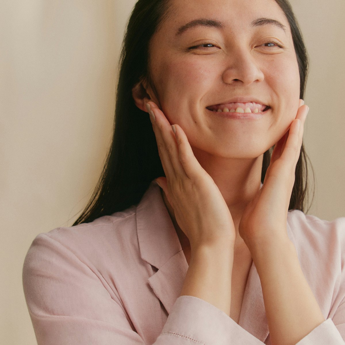 Photo showing a happy person smiling while putting on a face cream using both hands. Their long dark hair is brushed back behind their shoulders and they wear light rose pajamas.