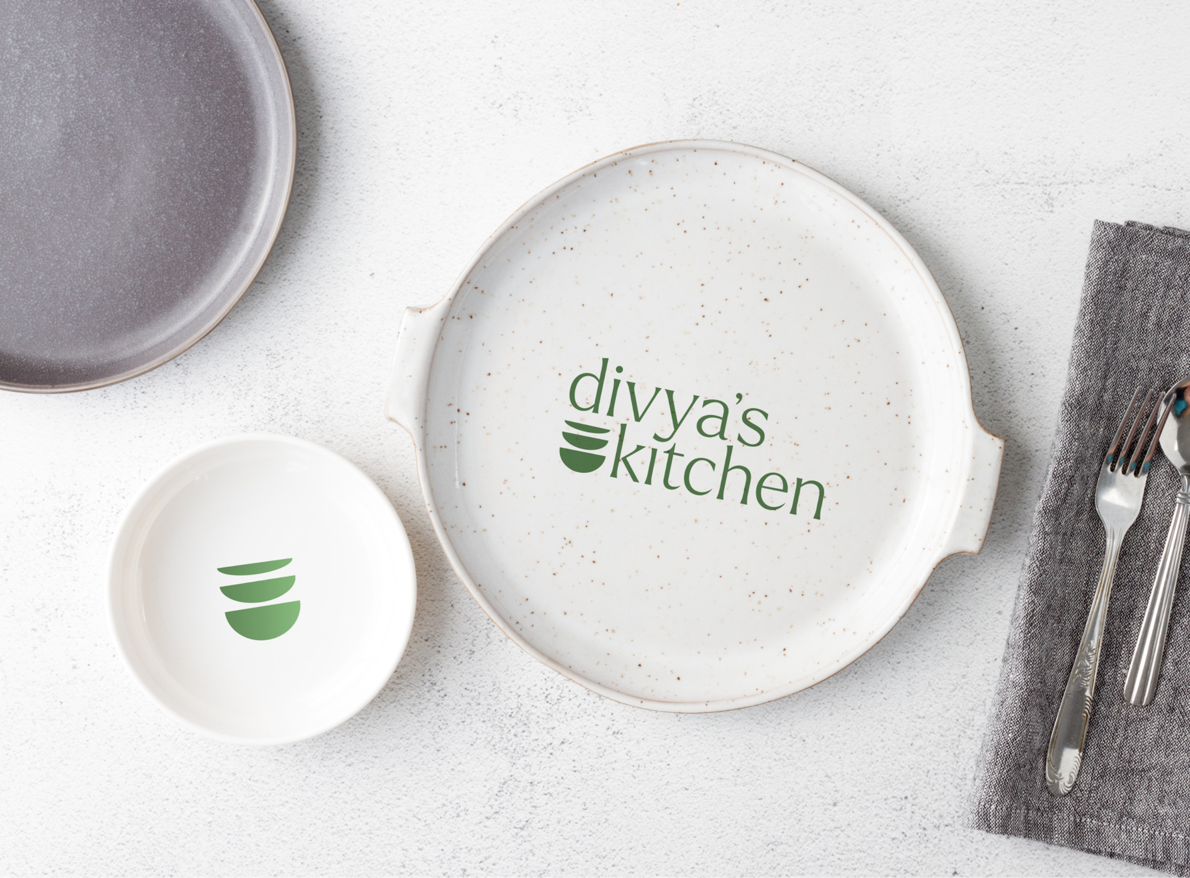 Three plates and silverwares on a white table. The one on top left is gray, the middle one is smallest with the bowls icon in the middle, and the right one is biggest and has the logo inside.