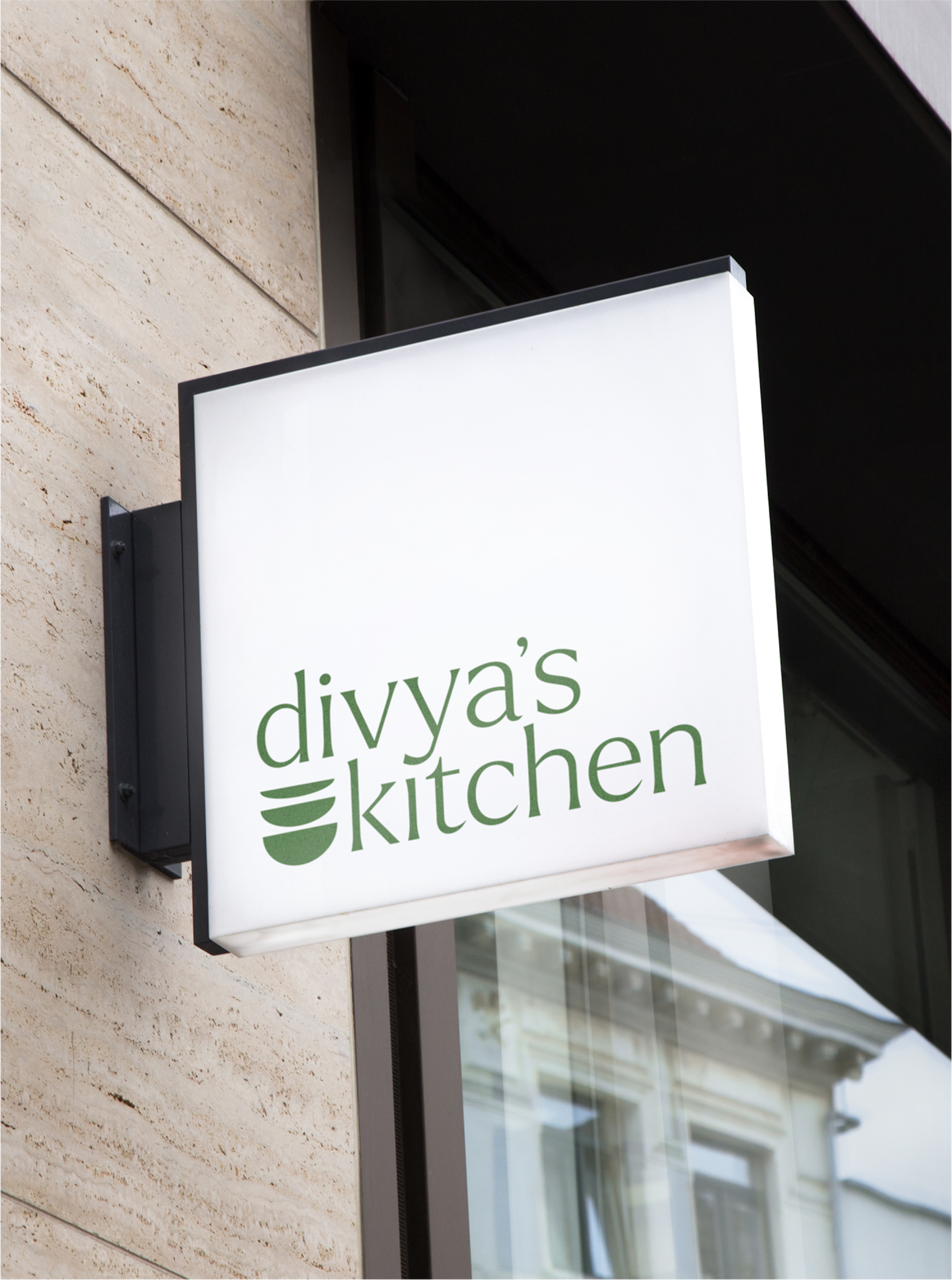 Divya's Signage with logo in green on a lower third of a white background. The logo is in a sharp serif font and it has 3 bowls icon next to the word "kitchen" on the second line.