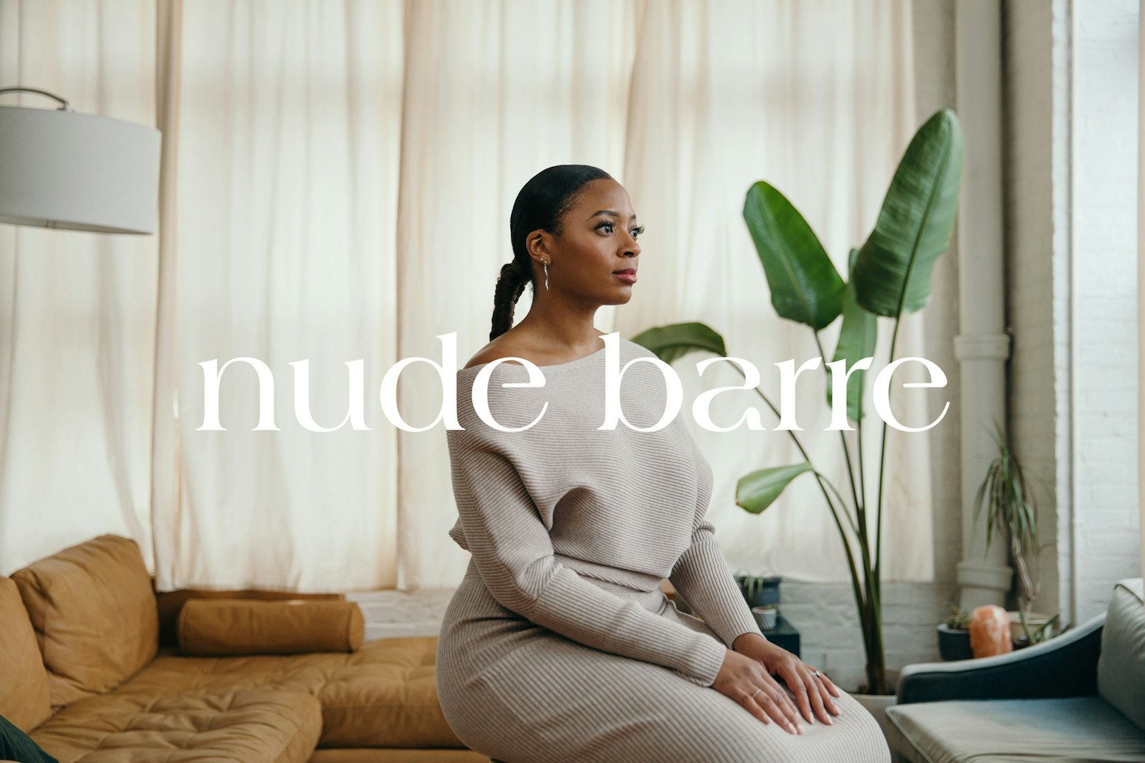 Image showing the founder of Nude Barre seated in their living room on the floor. They are wearing a knitted soft draping dress in a light warm gray color. Their hair is tied in a long braid down their back. Behind them is a cognac colored couch, a banana palm, and an oversized white lamp. The cream colors curtains in the background are closed and there is a salt-lamp in the far right corner.