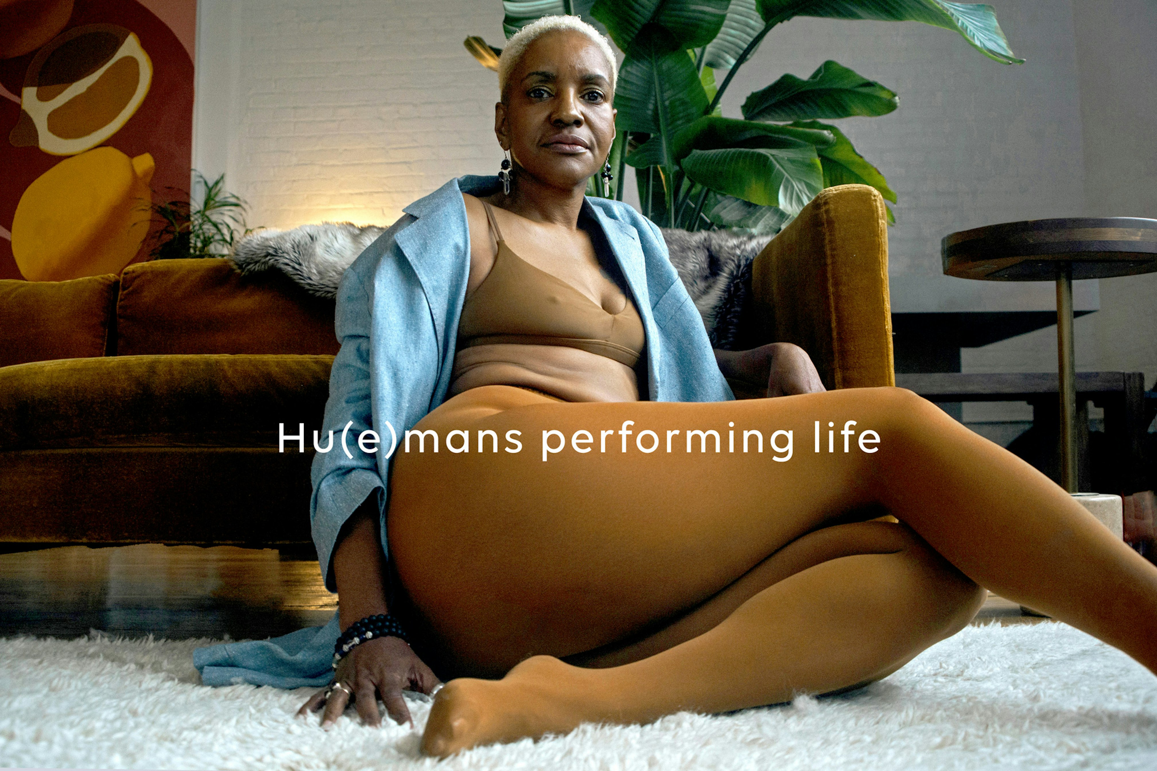 Image showing a person seated on a soft white rug against a mustard yellow velvet couch. Behind the couch is a big banana palm, a white brick wall, and a painting in warm colors. The person is looking in camera, has short white hair. They are wearing an open soft light blue denim shirt and underneath a Nude Barre bralette and tights. Across the image is written 'Hu(e)mans performing life'.