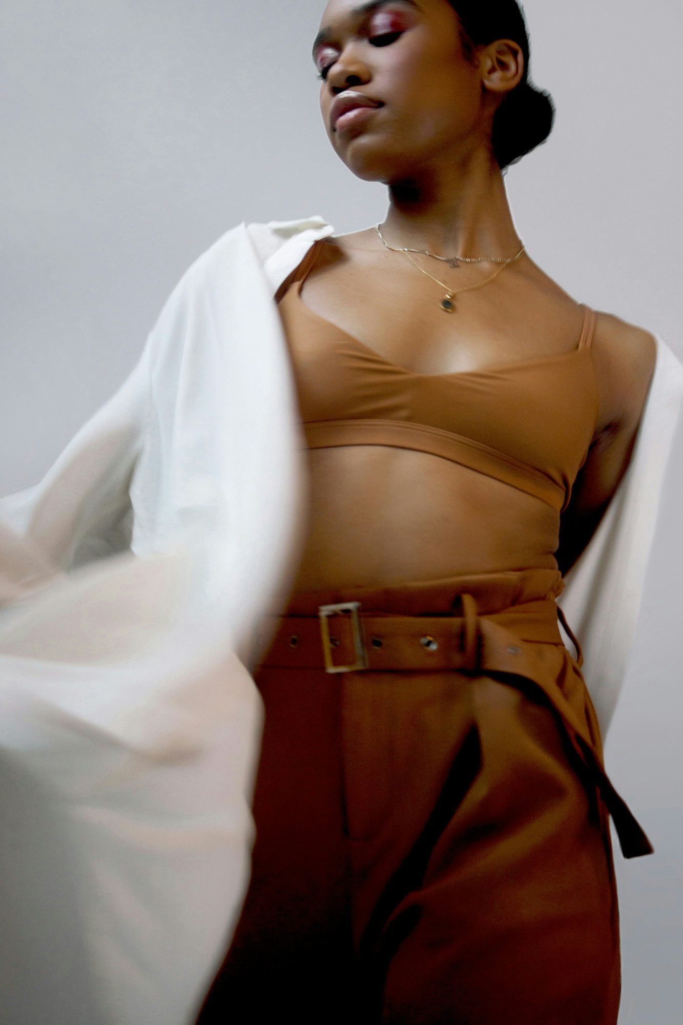 Cropped image showing person with their eyes closed standing against a gray wall. They are wearing a Nude Barre bralette and brown pants and an open white robe style shirt.
