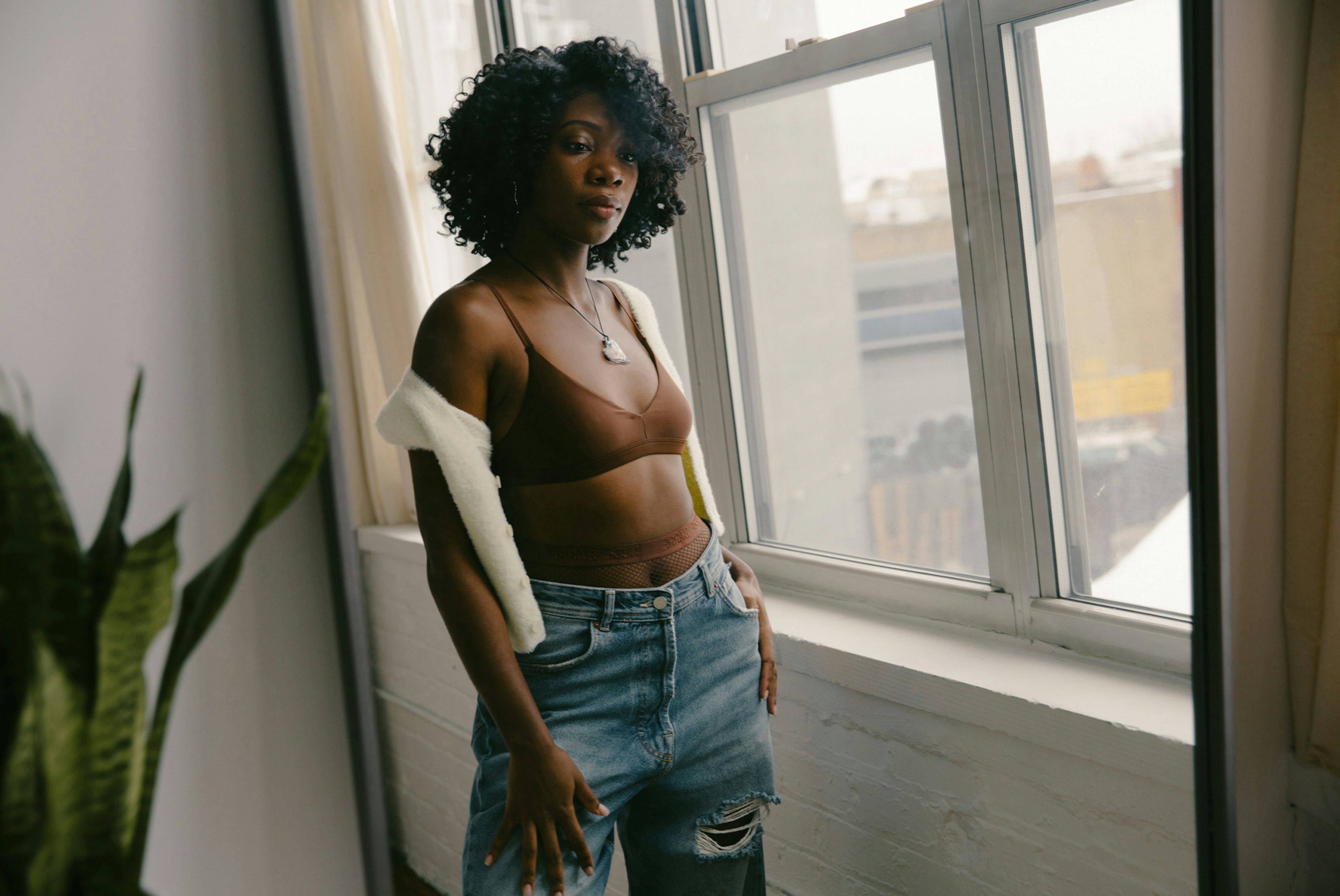 Image showing a person standing by a window, looking in camera. They are wearing a Nude Barre bralette and baggy mid blue jeans and a white short sleeve cardigan. Behind them is a green plant and the view of the window is a city.