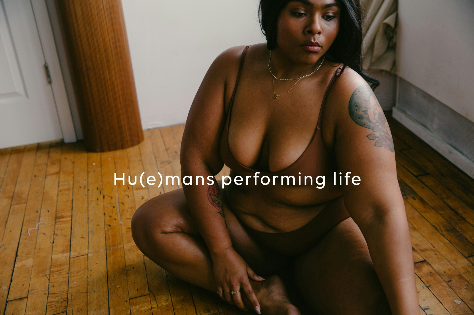 Cropped image showing a person sitting on a hard wood floor. They are wearing a Nude Barre bralette and underpants. They have a large tattoo on their left upper arm and their gaze is downwards. Across the image is written 'Hu(e)mans performing life'.