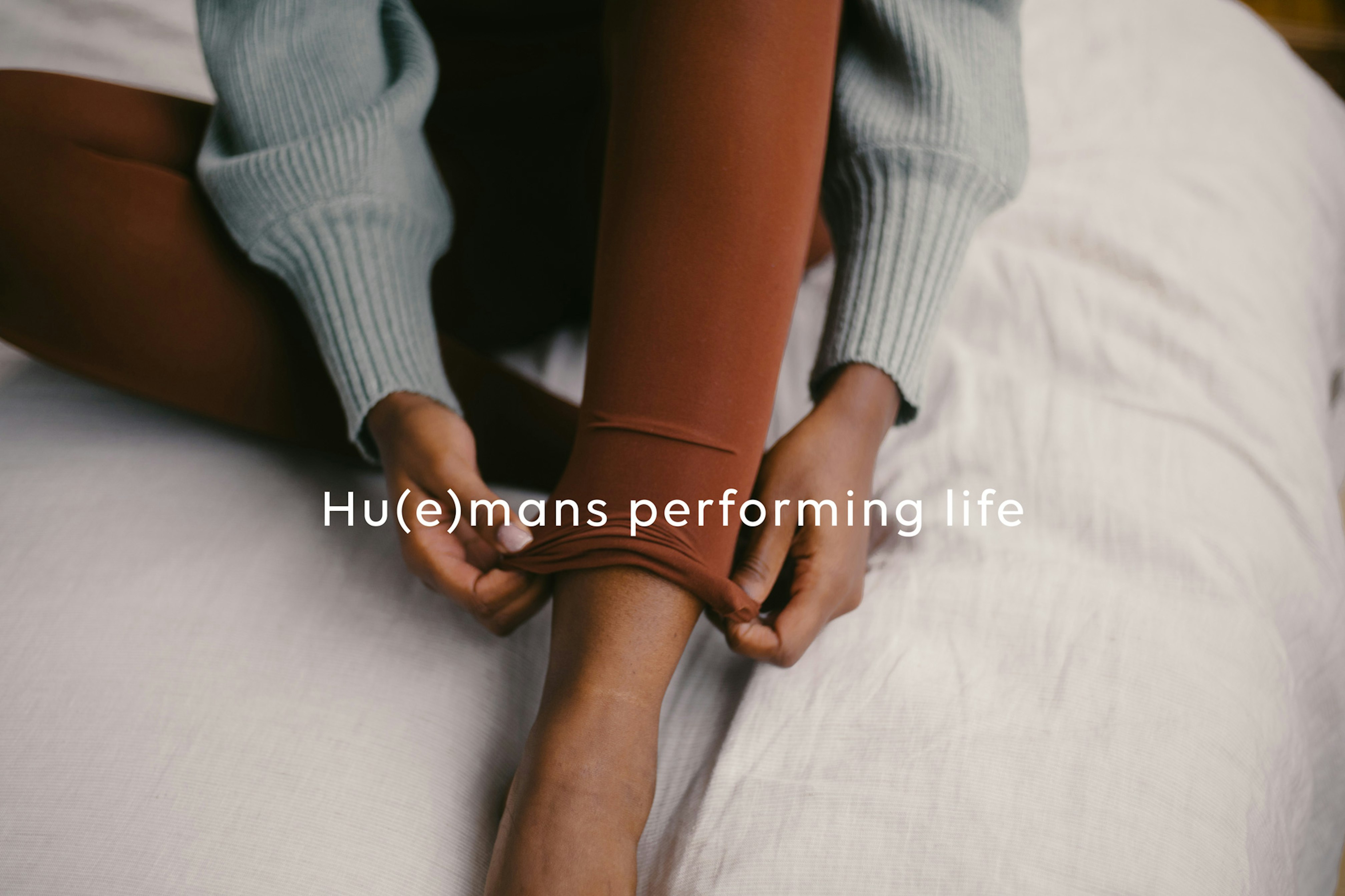 Close up image showing a pair of hands adjusting the bottom of a pair of leggings. The leggings are a rich brown and they are wearing a cool gray knitted sweater with long cuffs. They are sitting on what looks like a cream colored linen covered duvet. Across the image is written 'Hu(e)mans performing life'.