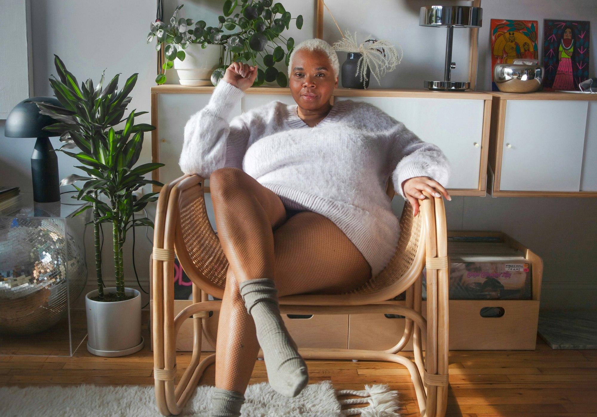 Image showing person sitting in a wicker chair, looking in camera, They are wearing a soft, oversized light gray sweater and medium warm gray socks. There are plants behind them and next to them and the sun is streaming in from the side.