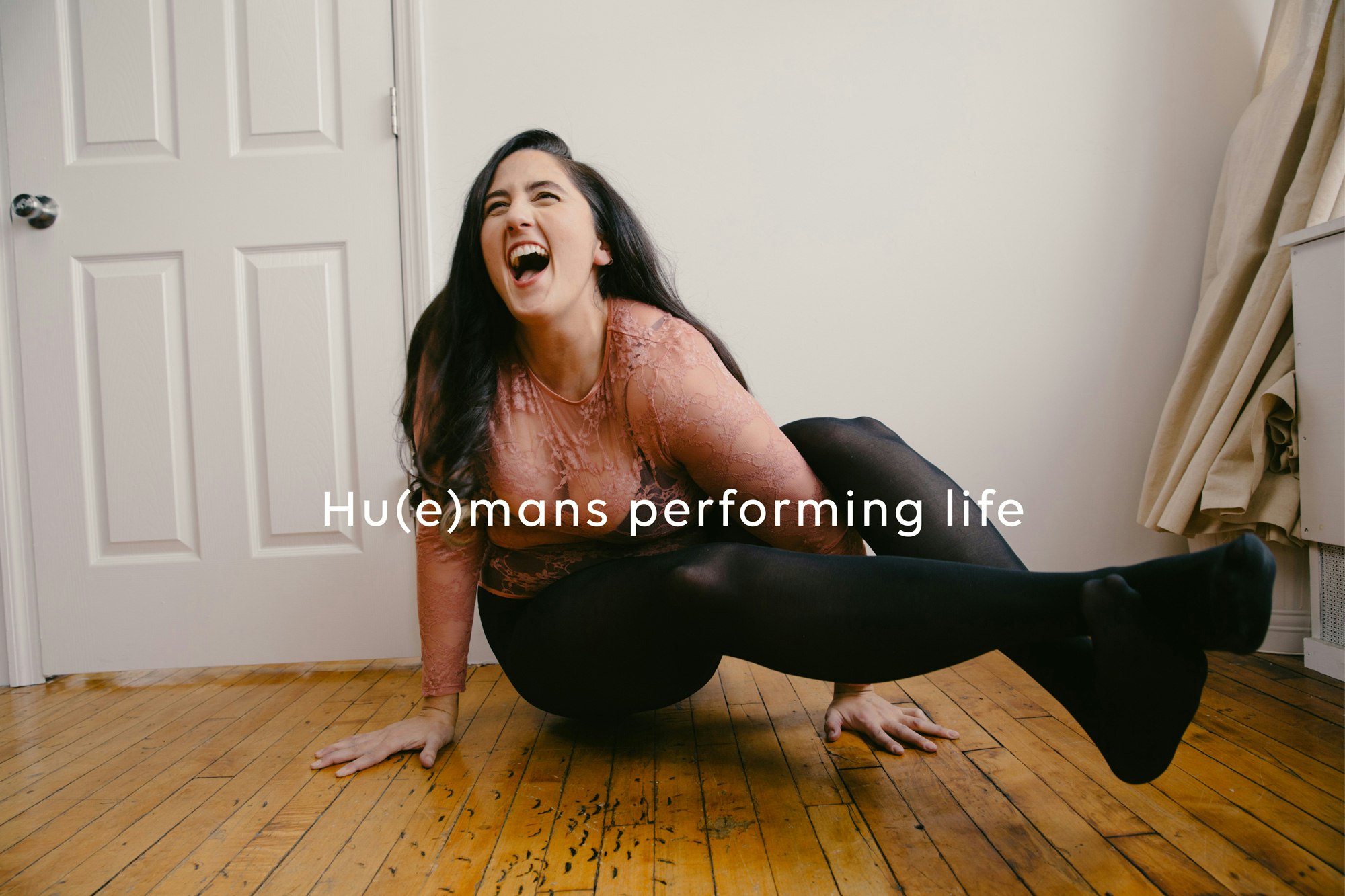 Image showing a person doing an advanced yoga pose (a type of crow pose it looks like). They are looking really happy, smiling, have long brown hair, is wearing a coral sheer top and black tights. The floor is a hardwood, medium brown, slim plank type. behind the person is a white wall and white door and next to them is what looks like a heavy canvas type cream colored curtain. Across the image it has the Nude Barre tagline "Hu(e)mans performing life".