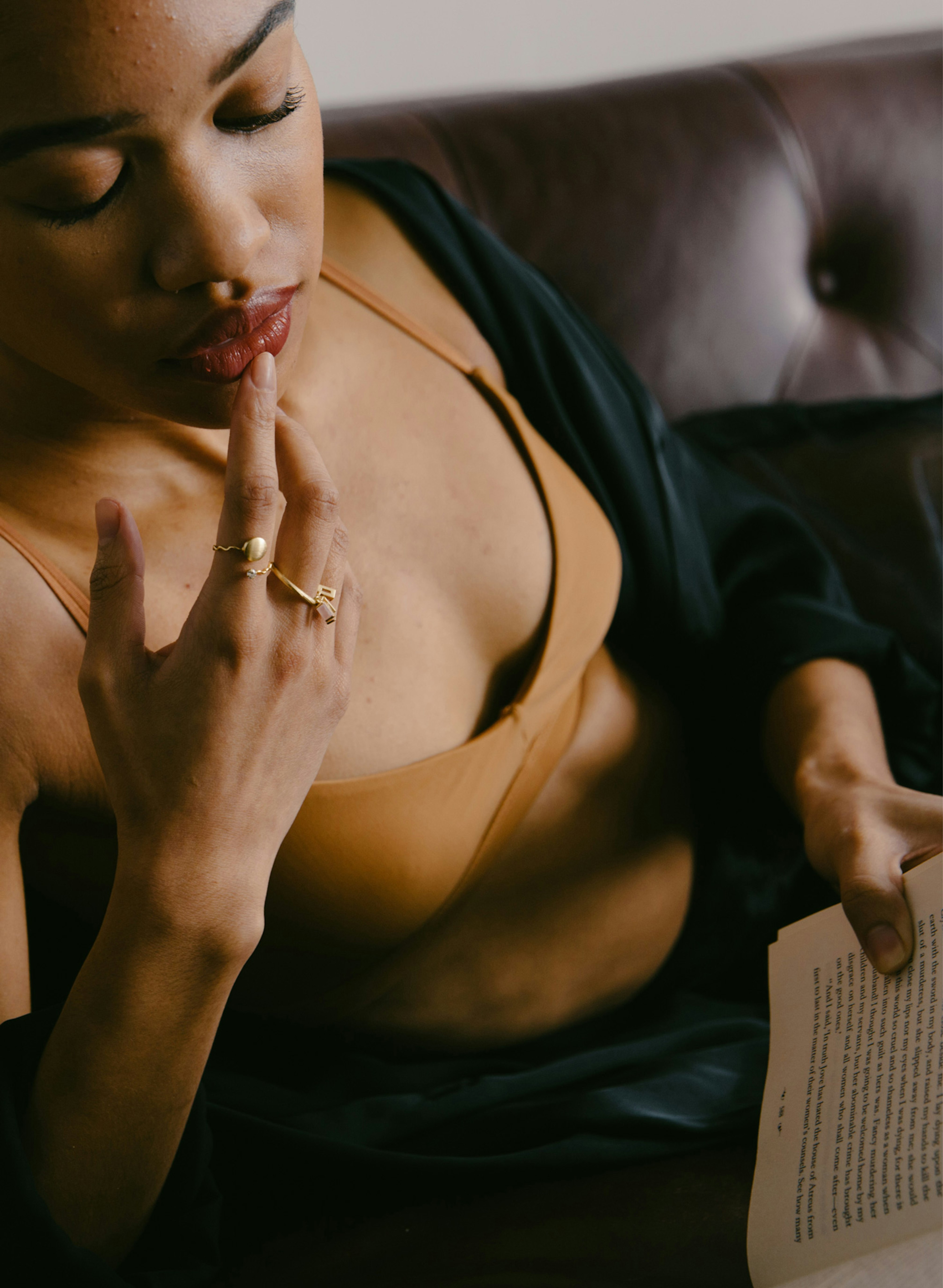 Close up image showing a person wearing a Nude Barre bralette and reading a book. They are holding the book with their left hand while softly touching their mouth with the right hand. They are sitting on a dark brown couch.