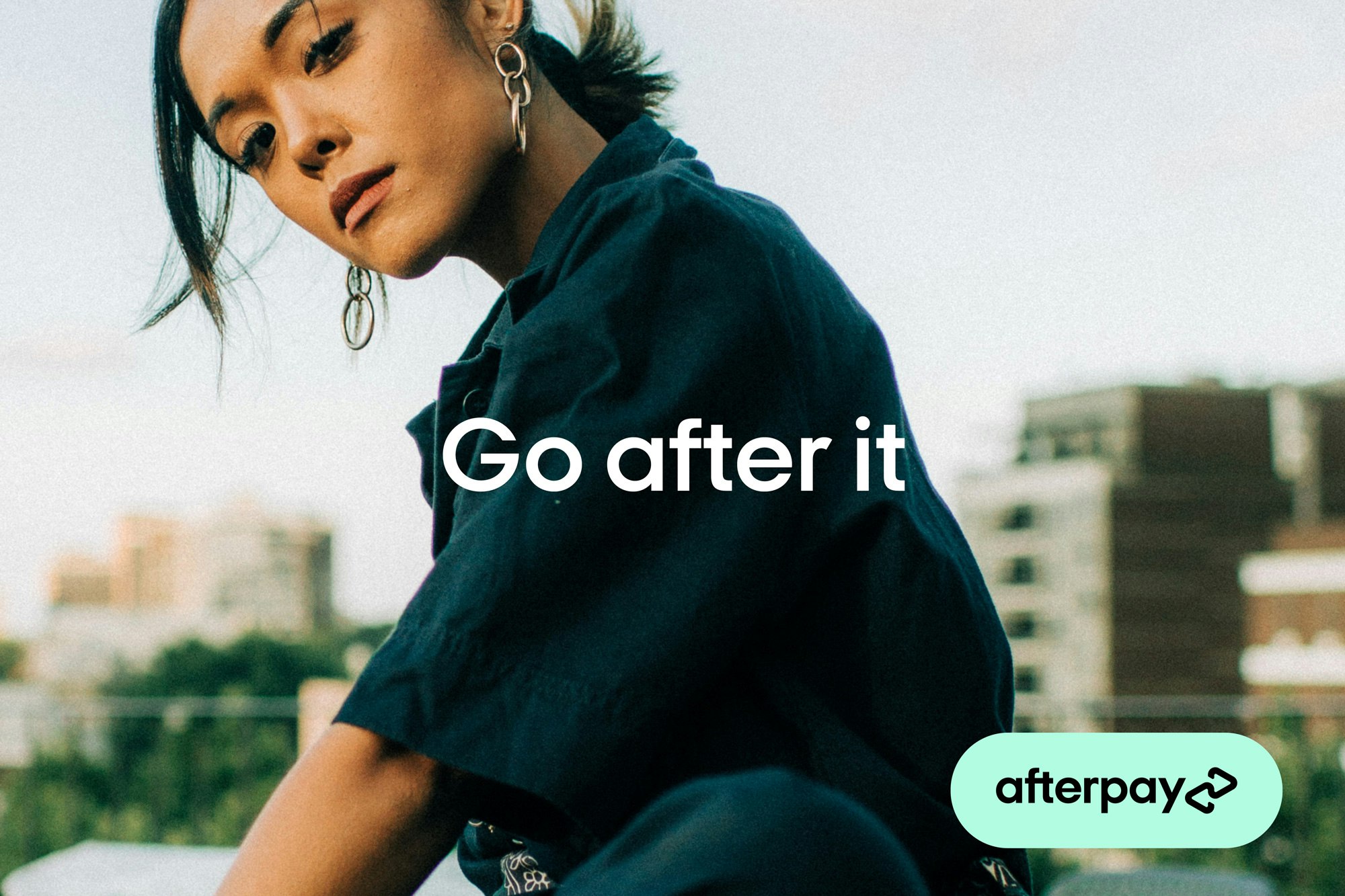 Close up image showing person looking in camera. They are wearing a dark teal short sleeved shirt, silver earrings and in the background there are buildings of what looks like a cityscape. Across the image it says GO AFTER IT and in the bottom right corner there is a Bondi mint pill saying Afterpay inside in black.