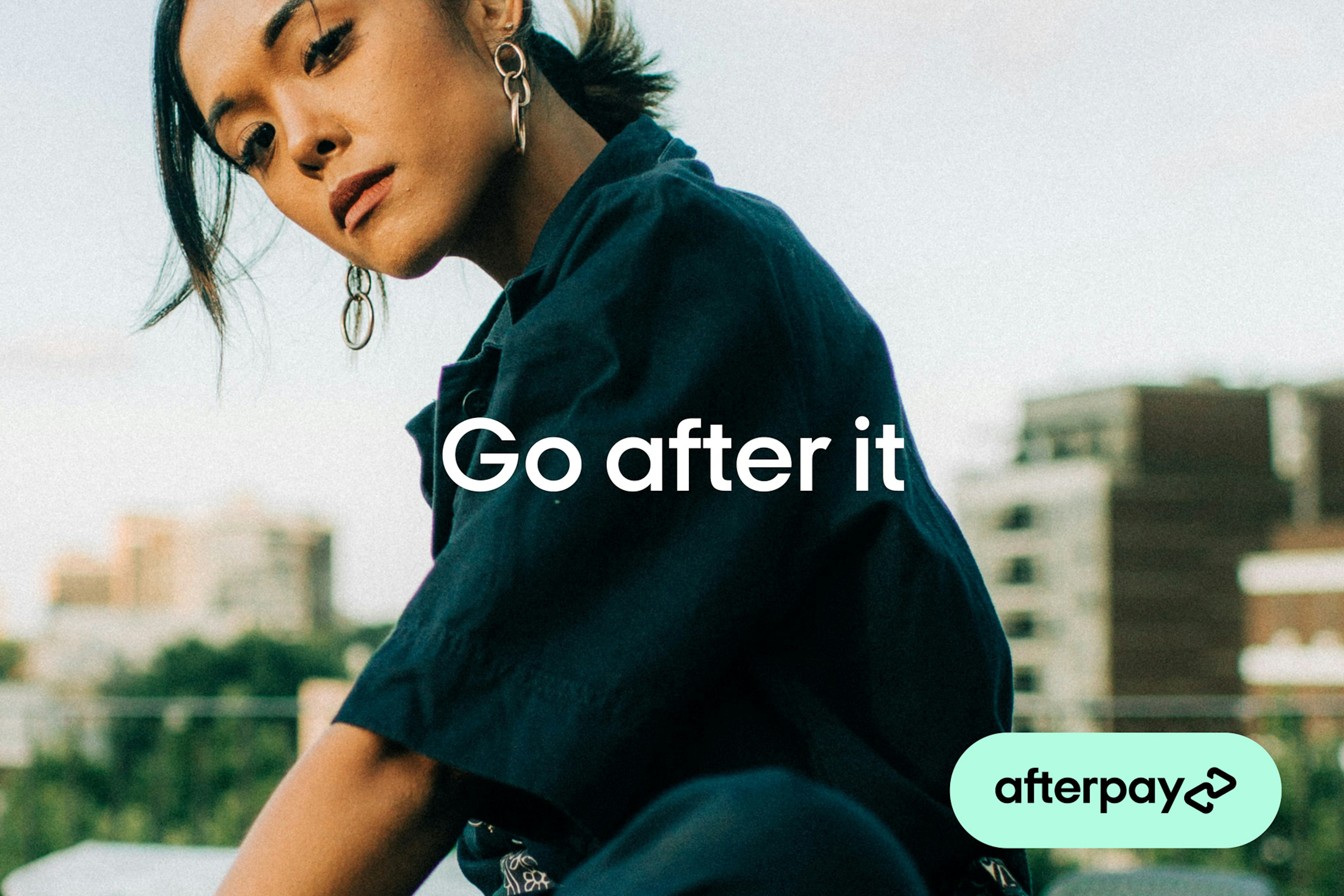 Close up image showing person looking in camera. They are wearing a dark teal short sleeved shirt, silver earrings and in the background there are buildings of what looks like a cityscape. Across the image it says GO AFTER IT and in the bottom right corner there is a Bondi mint pill saying Afterpay inside in black.
