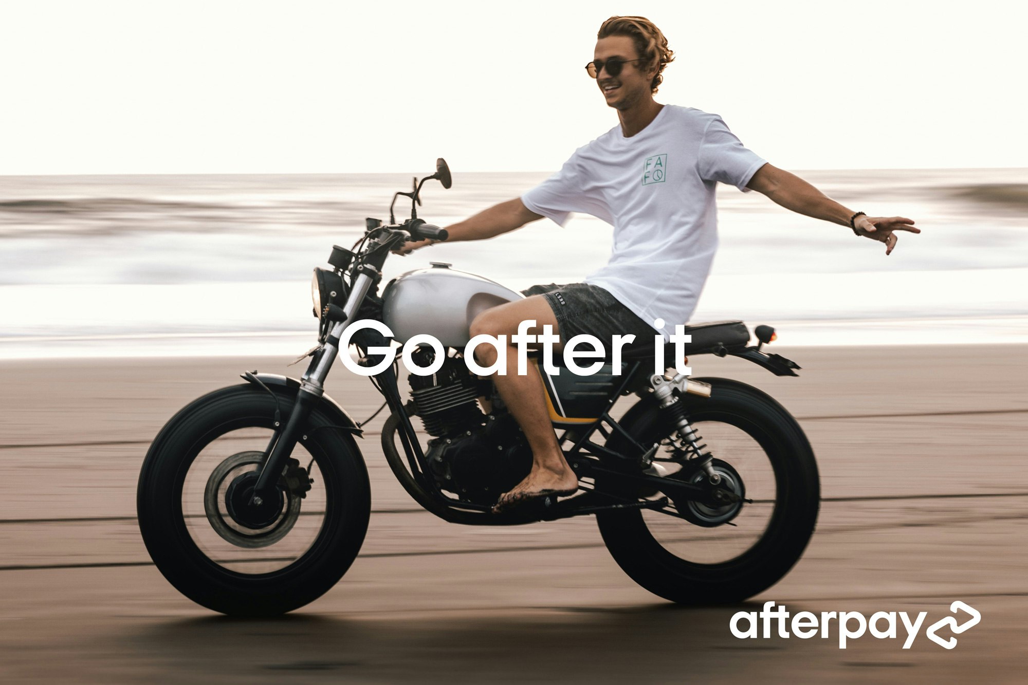 Image showing a person riding a motorcycle on the beach. They look very delighted and is steering with only the right hand while the left arm is floating out in the air. They are wearing sunglasses, a white teeshirt, gray shorts and have bare feet. The motorcycle looks vintage, the sand looks wet and flat, and the sea behind them is out of focus. Across the image it says GO AFTER IT and in the bottom right corner there is Afterpay logo in white.