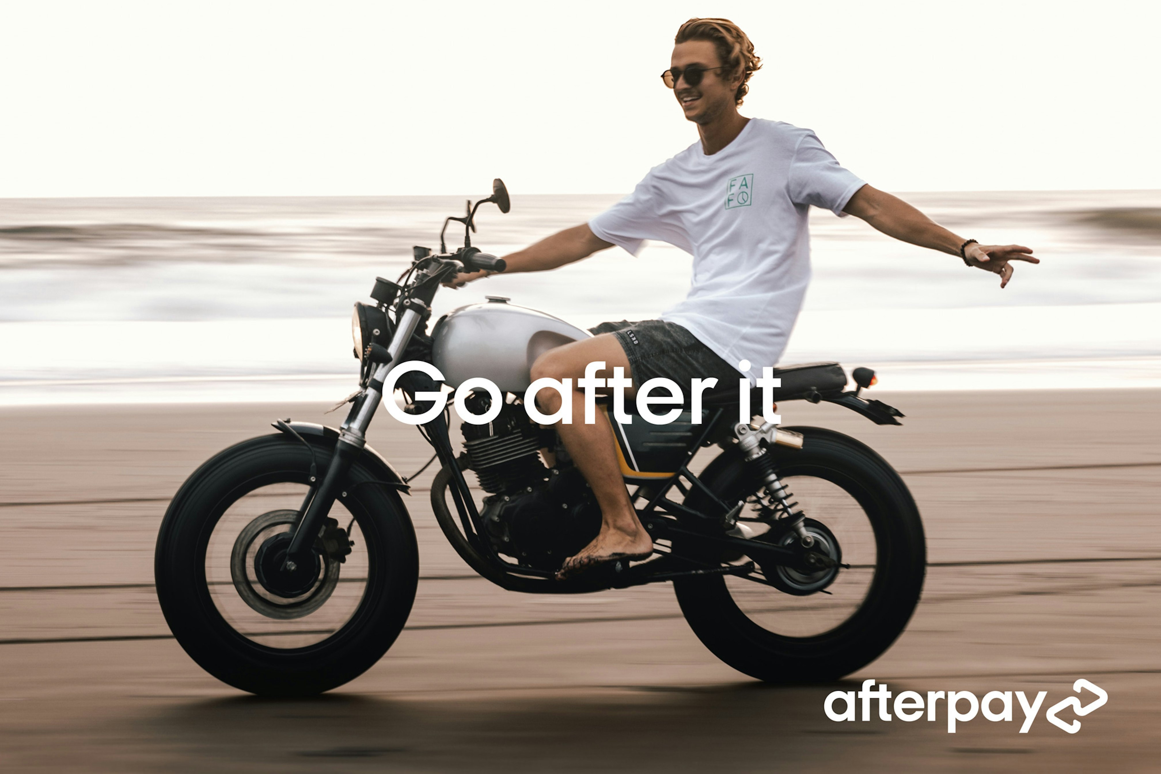 Image showing a person riding a motorcycle on the beach. They look very delighted and is steering with only the right hand while the left arm is floating out in the air. They are wearing sunglasses, a white teeshirt, gray shorts and have bare feet. The motorcycle looks vintage, the sand looks wet and flat, and the sea behind them is out of focus. Across the image it says GO AFTER IT and in the bottom right corner there is Afterpay logo in white.