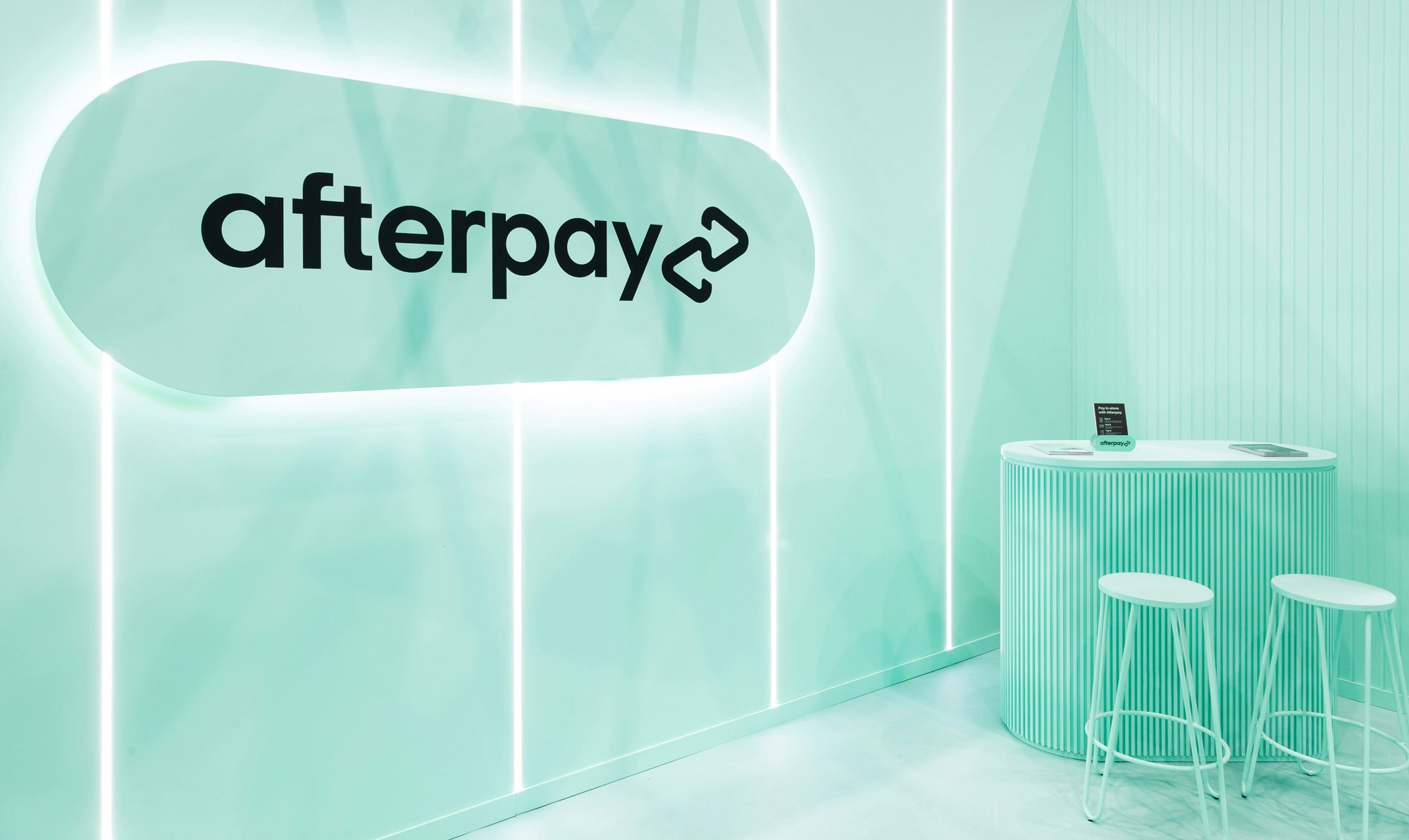 Image showing the interior of the Afterpay expo booth. Everything is Bondi Mint and there is an oversized logo pill which is backlit. In the back is a mint table and two mint stools. On top of the table are black pamphlets describing Afterpay.