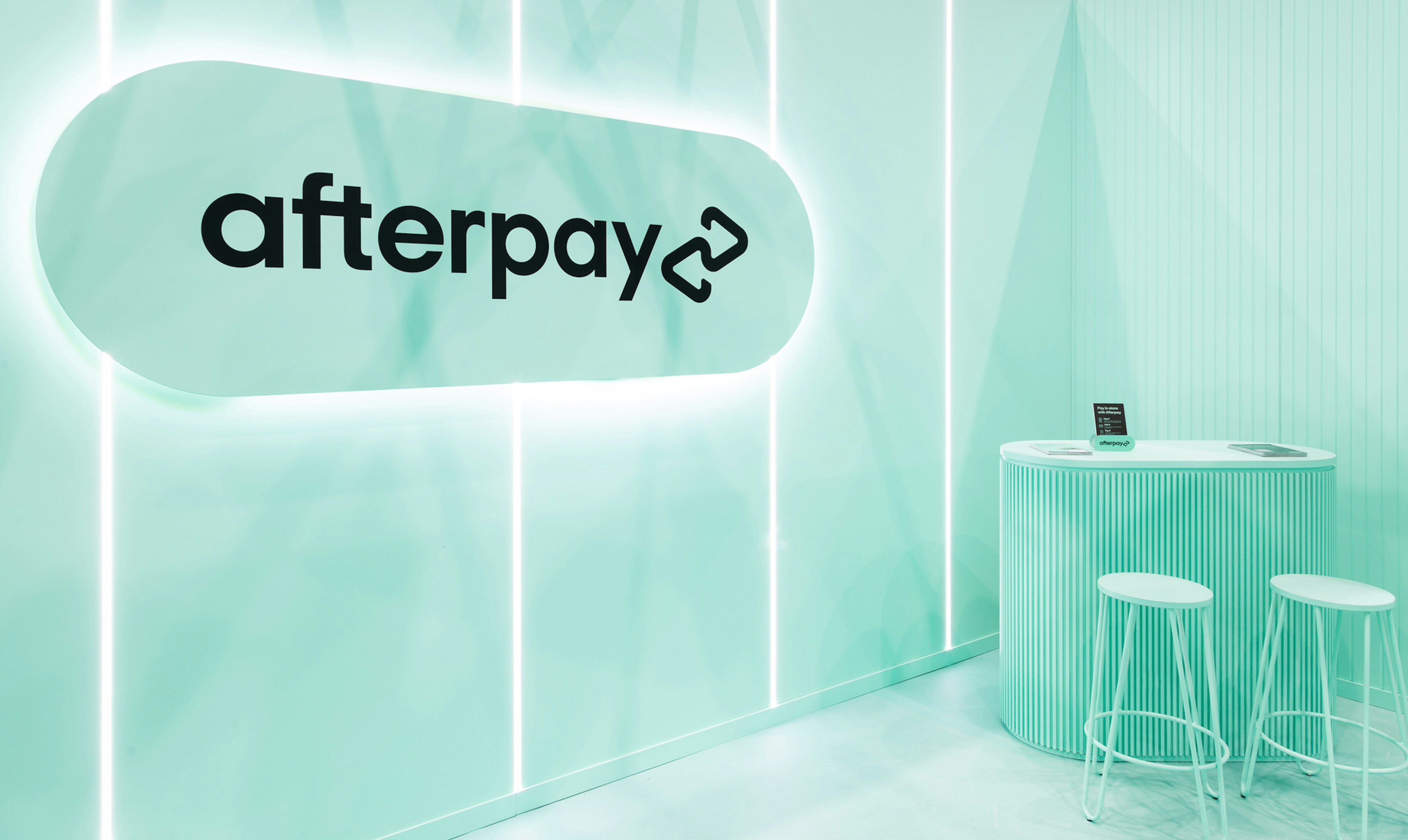 Image showing the interior of the Afterpay expo booth. Everything is Bondi Mint and there is an oversized logo pill which is backlit. In the back is a mint table and two mint stools. On top of the table are black pamphlets describing Afterpay.