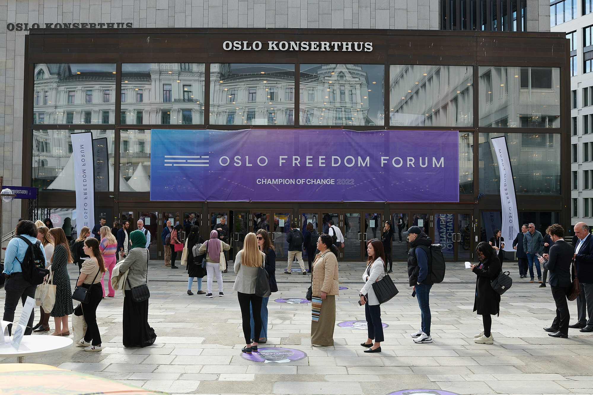 Front of Oslo Konserthus with a banner saying Oslo Freedom Forum Champion of Change 2022 in white on a purple blue gradient background. People are in line in front of it.