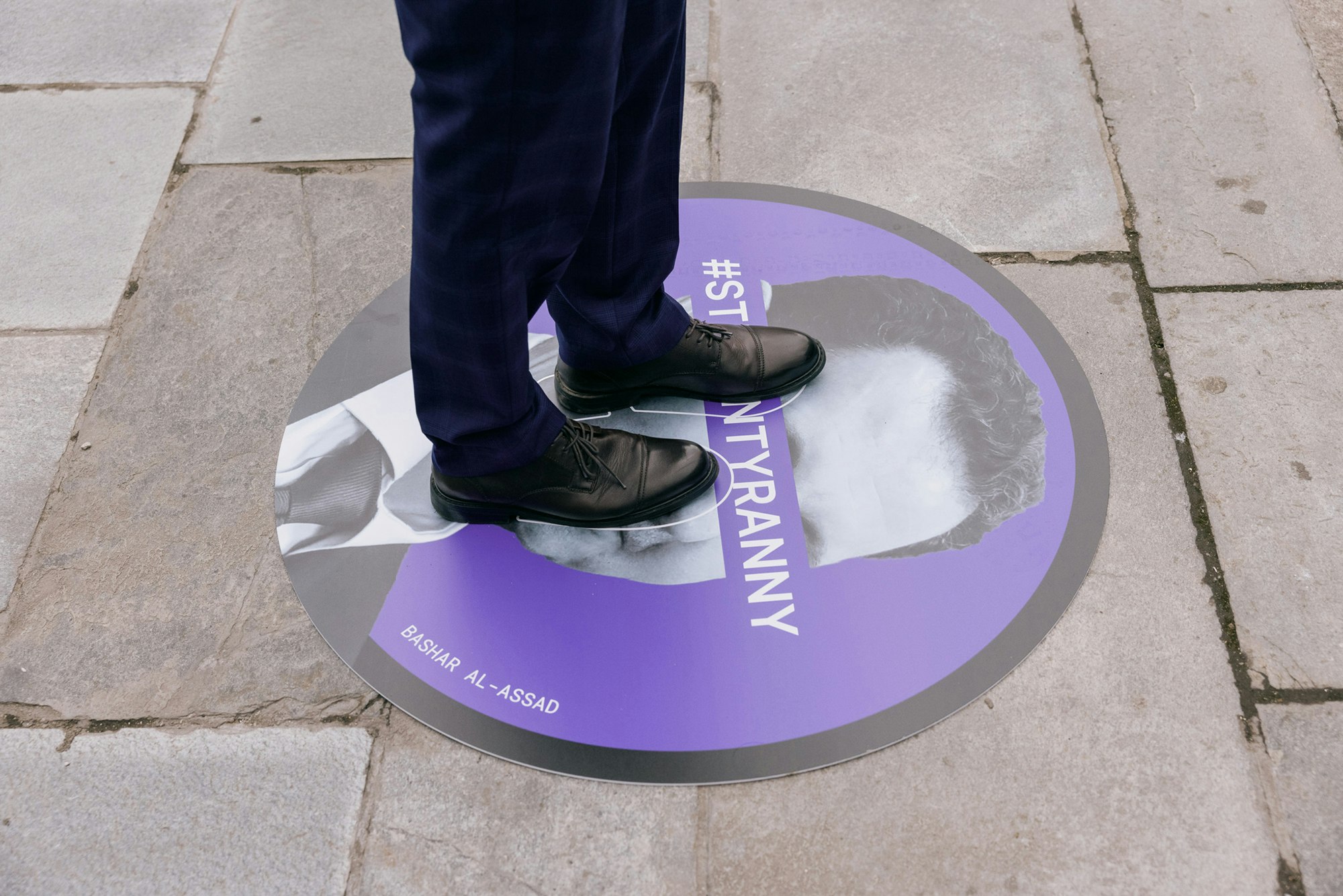 A person stepping on a circle mat that has a picture of Bashar Al-Assad in black and white on a purple background with thick black stoke with a purple banner saying #StepOnTyranny across his eyes.