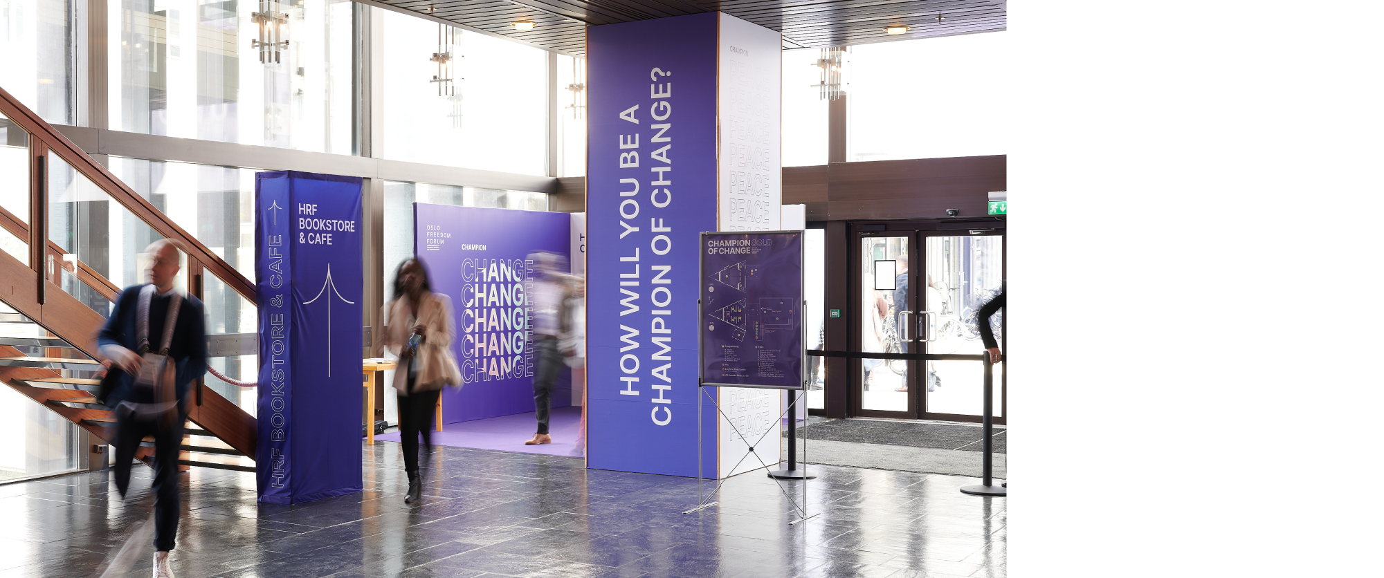 Interior of the Konserthus. There's testimonial stands in the corner. In front of it is lobby column saying "How will you be a champion of change?" in white on a purple background. Next to it is a hop up in front of the stairs saying HRF Bookstore & Cafe with an arrow pointing up. There're people walking in between.