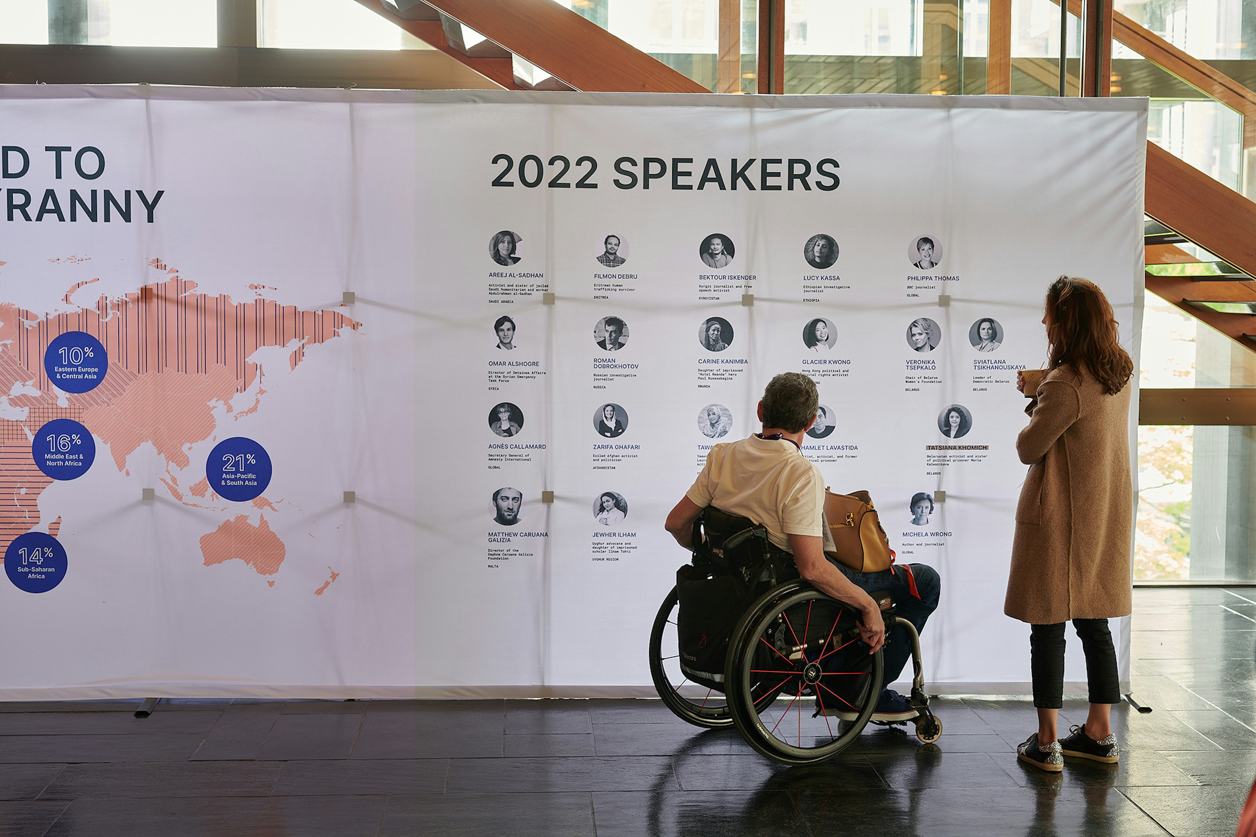 There're two people looking at a hop up with the speakers information. The person on the left is on a wheel chair with a purse on his lap. The person on the right is wearing a long coat and holding a paper cup.