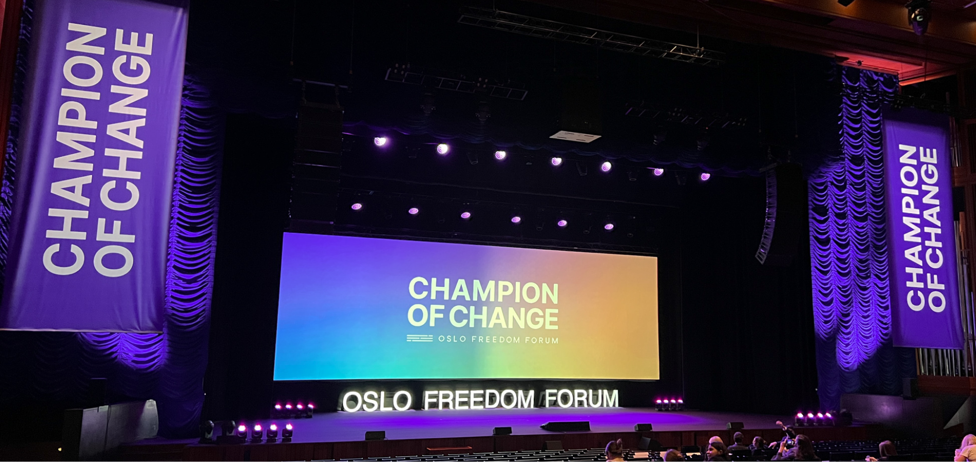 The theatre stage of the konserthus. On a screen is Champion of Change logo in white in a brand gradient. Underneath is dimensional letter saying Oslo Freedom Forum. On the two sides, there're purple banners saying Champion of change in white.