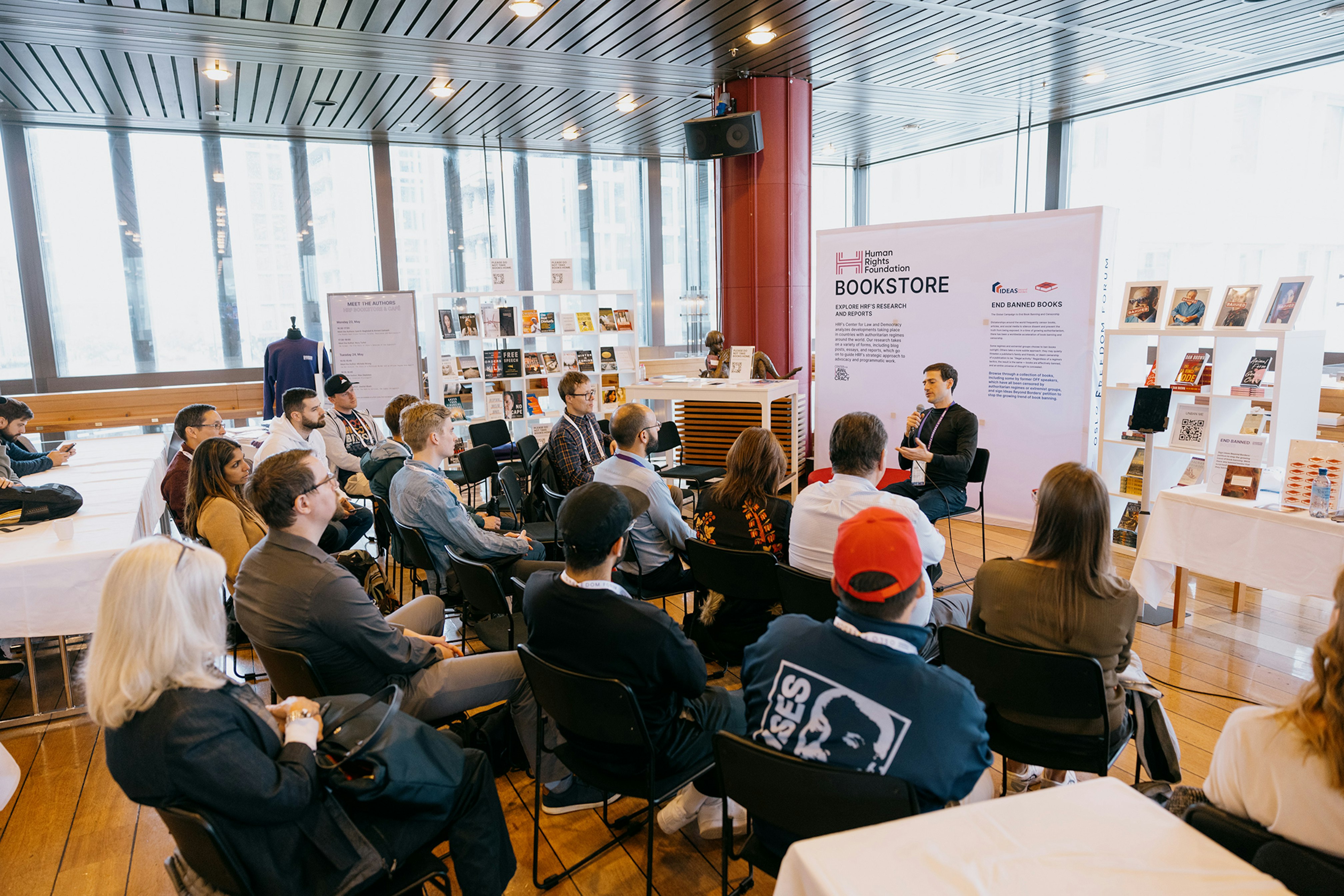 A person talking in front of an audience. Behind is a hop up for HRF Bookstore.