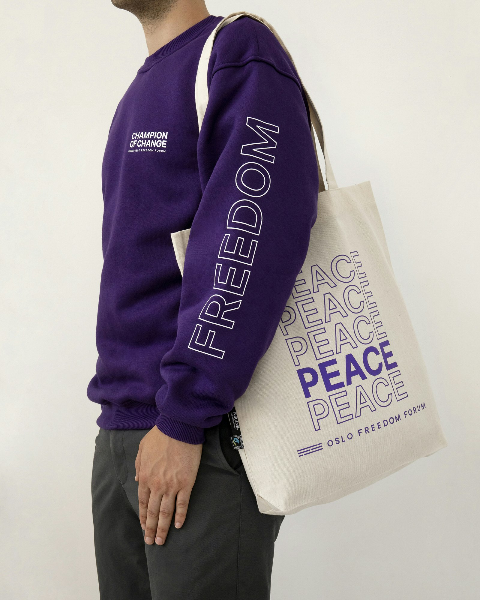 A person in a purple sweatshirt with Champion of change logo in white on top right of the shirt and a word saying "Freedom" in white stokes. He's also having a tote bag saying peace repeatedly and Oslo Freedom Forum logo at the bottom.