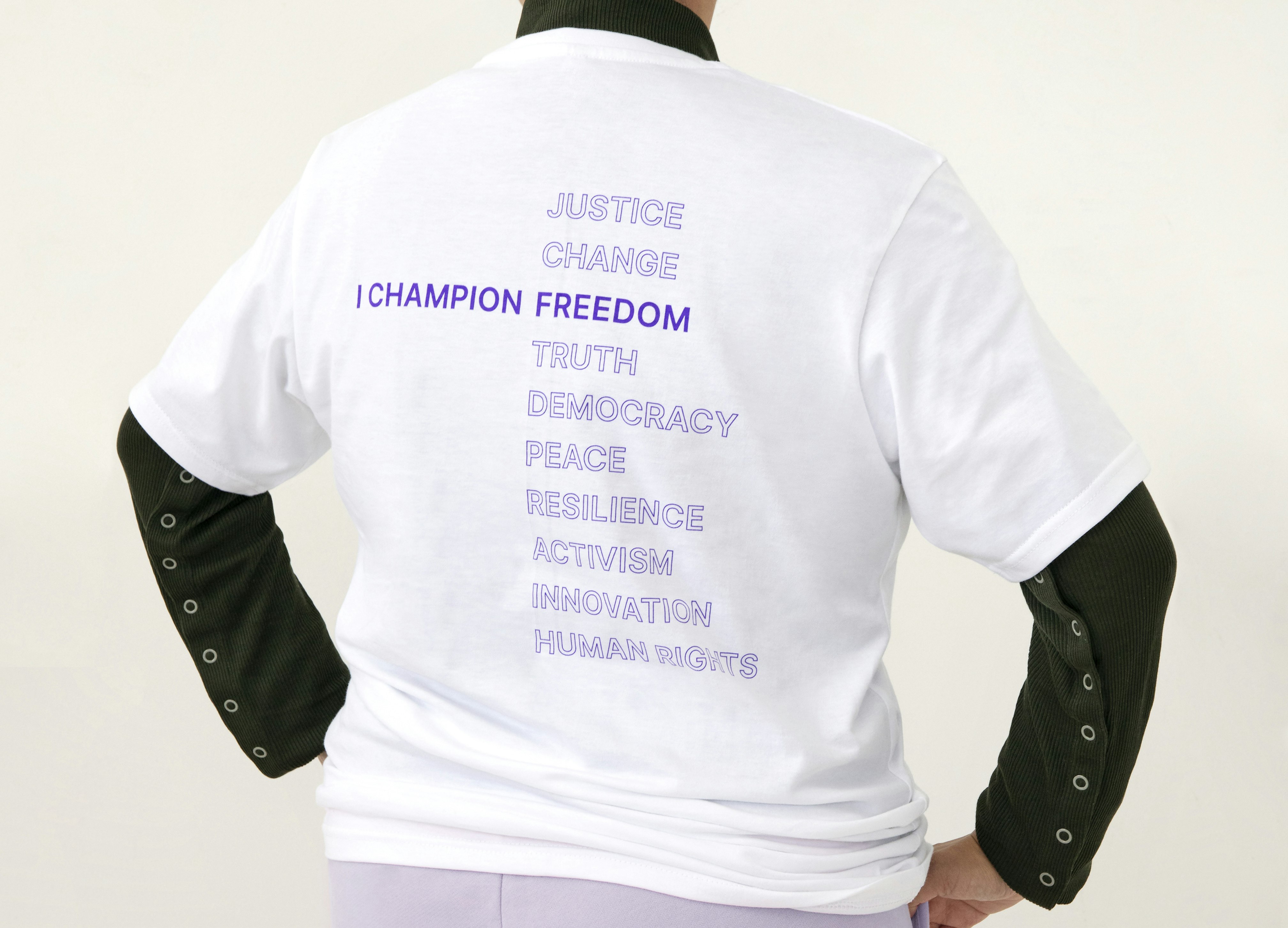 A person wearing short sleeves white shirt saying I Champion Freedom in purple and the words "Justice, Change, Truth, Democracy, Peace, Resilience, Activism, Innovation, Human Rights" aligning with Freedom in purple strokes.